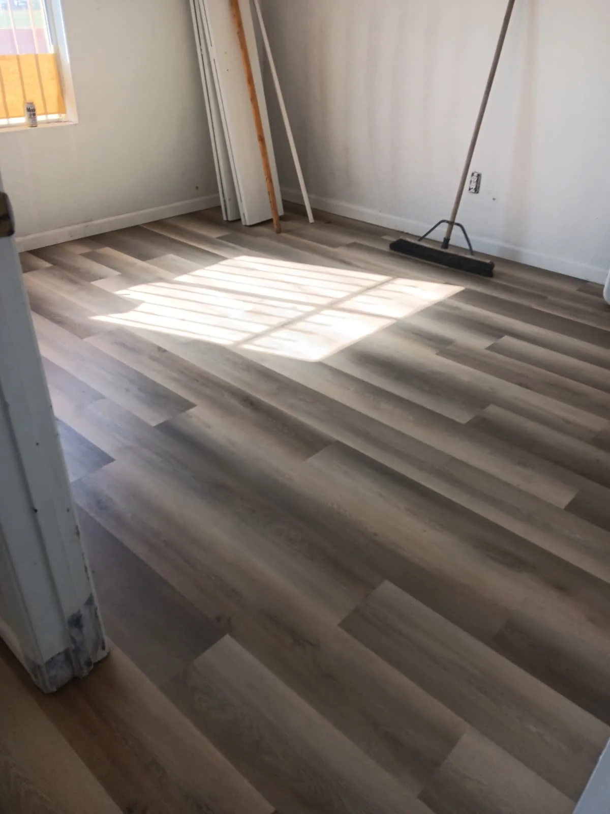 Room with newly installed light wood laminate flooring, sunlight casting shadows through a window, with construction tools and materials leaning against the wall.