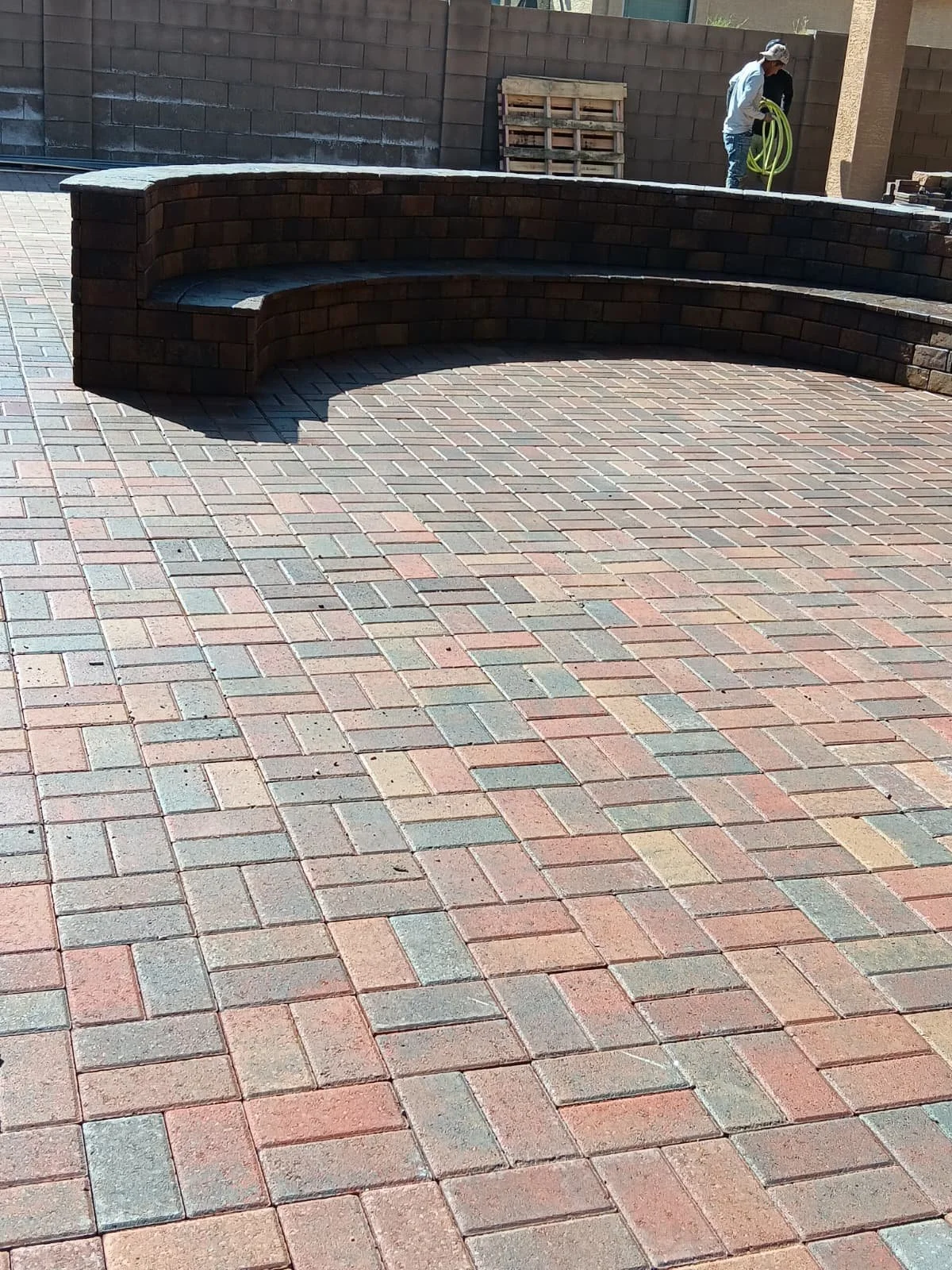 A curved brick seating area in an outdoor space with colorful brick pavers on the ground, and a worker in the background holding a yellow hose.