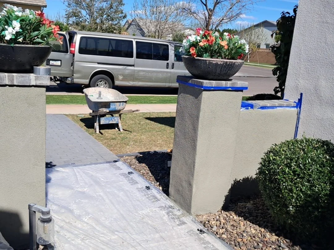 Front yard with two large flower pots with colorful flowers on concrete pillars, a wheelbarrow on grass, parked van on street, clear blue sky, and a well-maintained bush.