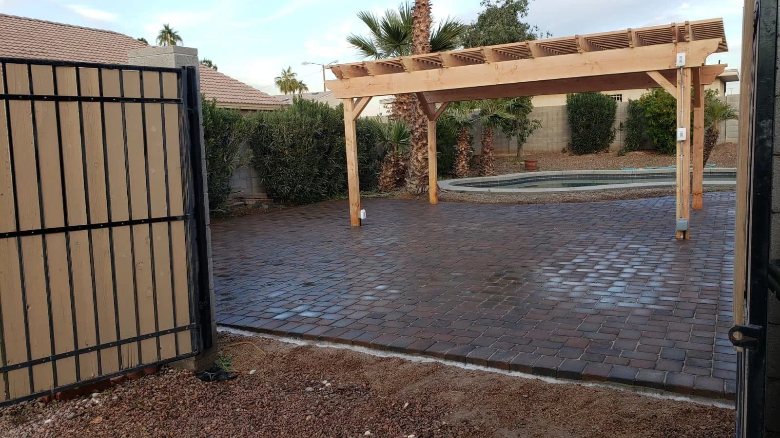 Backyard patio with newly installed brick pavers, a wooden pergola, a swimming pool in the background, and desert bushes and palm trees.