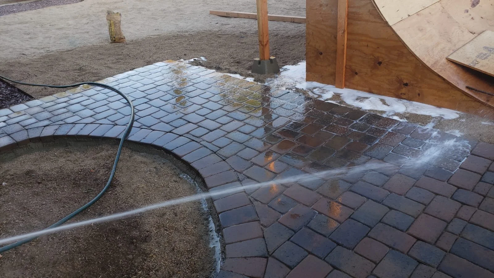 Outdoor scene of new brick pathway being washed with a garden hose, with construction materials and wooden structure in the background.