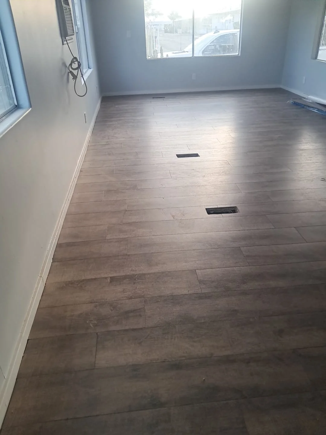 Empty room with large windows, new light brown wood-style flooring, and two floor-level air vents.