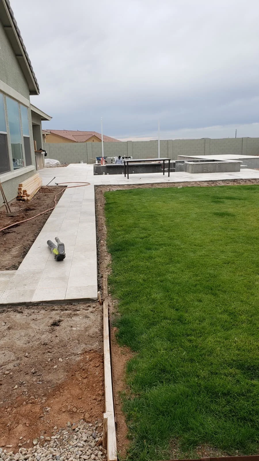 Backyard under construction with partially completed patio, a grassy lawn, construction tools, and equipment, with a gray sky overhead.