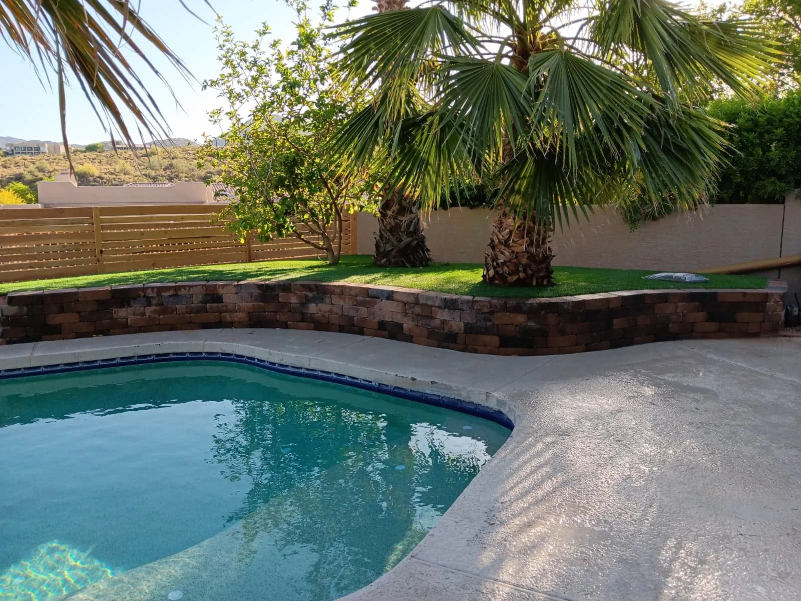 Residential backyard with a swimming pool, palm trees, a grassy area, a wooden fence, and a beige wall, with hills in the background.