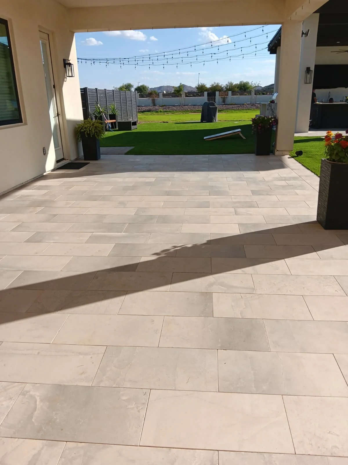 View of a backyard patio with beige tiles, potted plants, string lights, artificial grass, and a cornhole game, with a fenced yard and blue sky in the background.