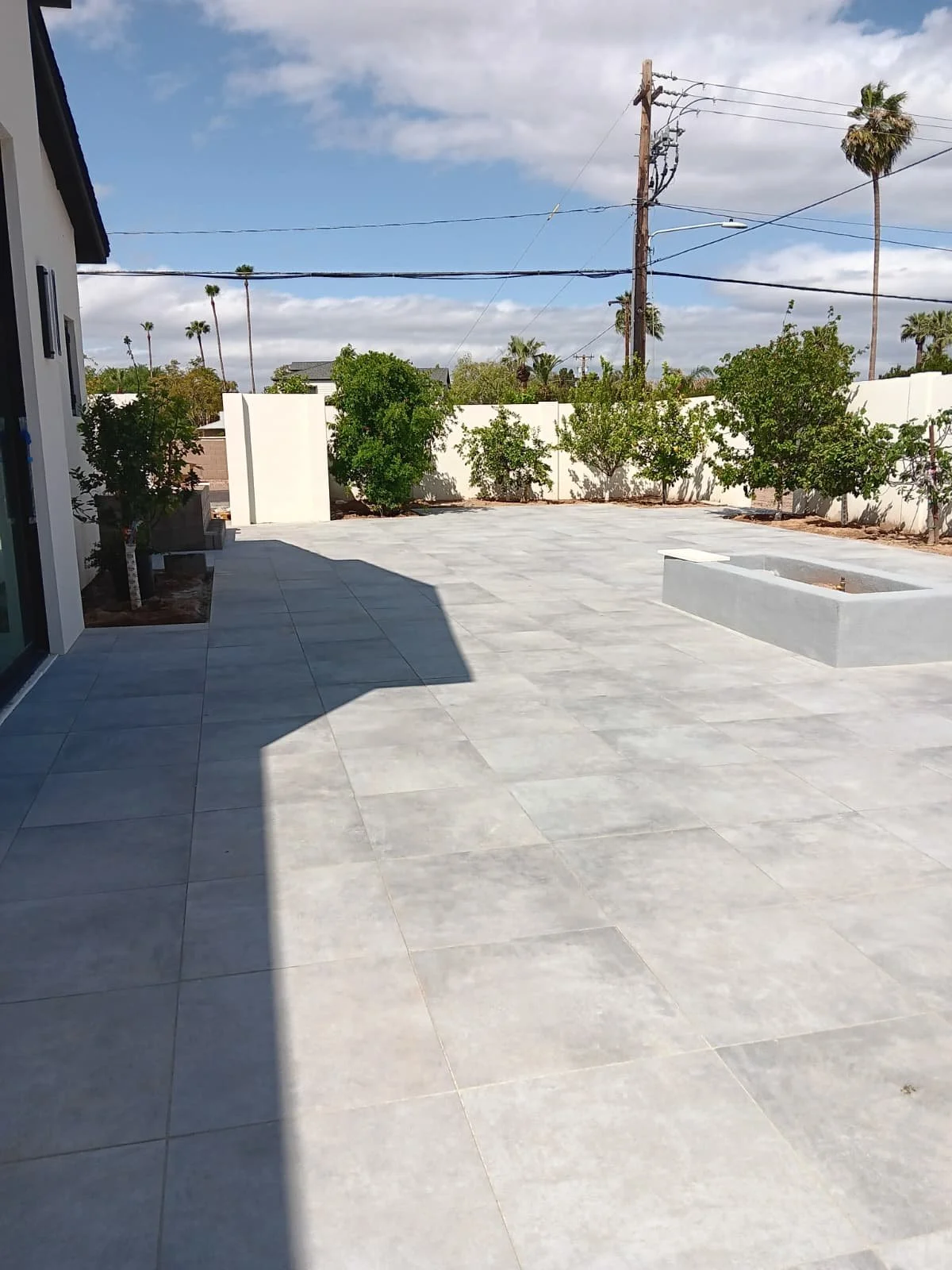 A modern backyard patio with large gray tiles, green trees and shrubs along a white fence, palm trees in the background, and utility poles with power lines under a partly cloudy sky.