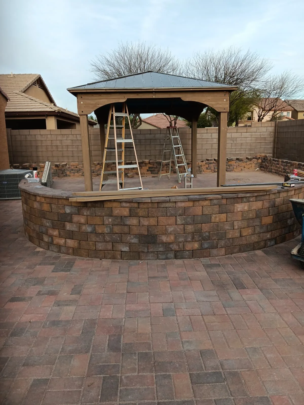 A backyard patio with a brick fire pit and a wooden gazebo under construction, with ladders inside.