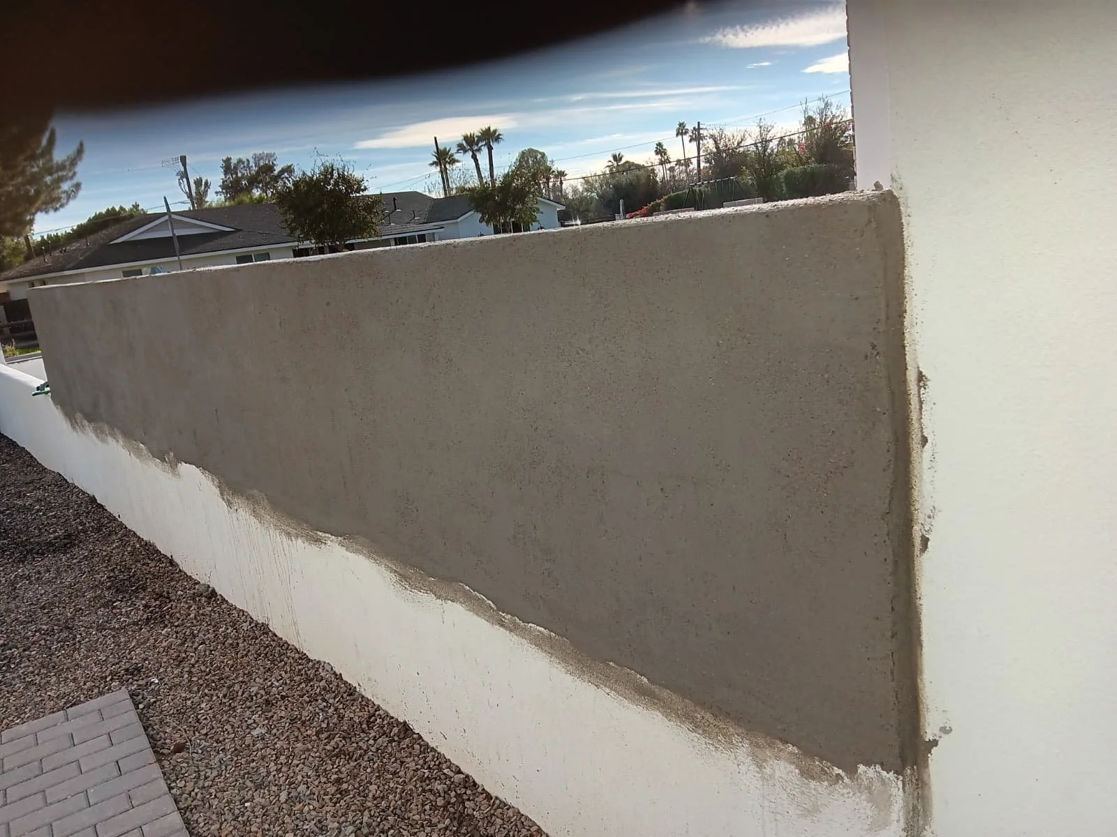 Close-up view of a freshly coated white concrete wall with a beige top layer, with a gravel pathway and a small section of paver stones in the foreground. Residential houses and palm trees are visible in the background under a partly cloudy sky.