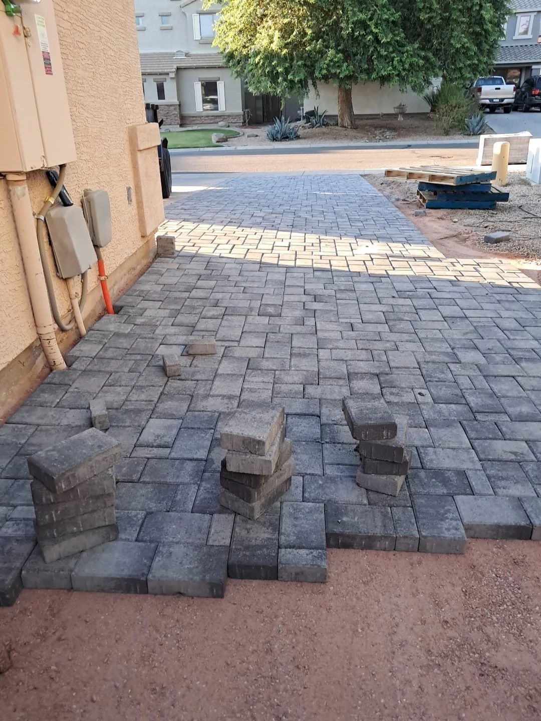 Paved pathway under construction with stacks of bricks in the foreground, a tree and neighborhood houses in the background, and construction materials to the right.