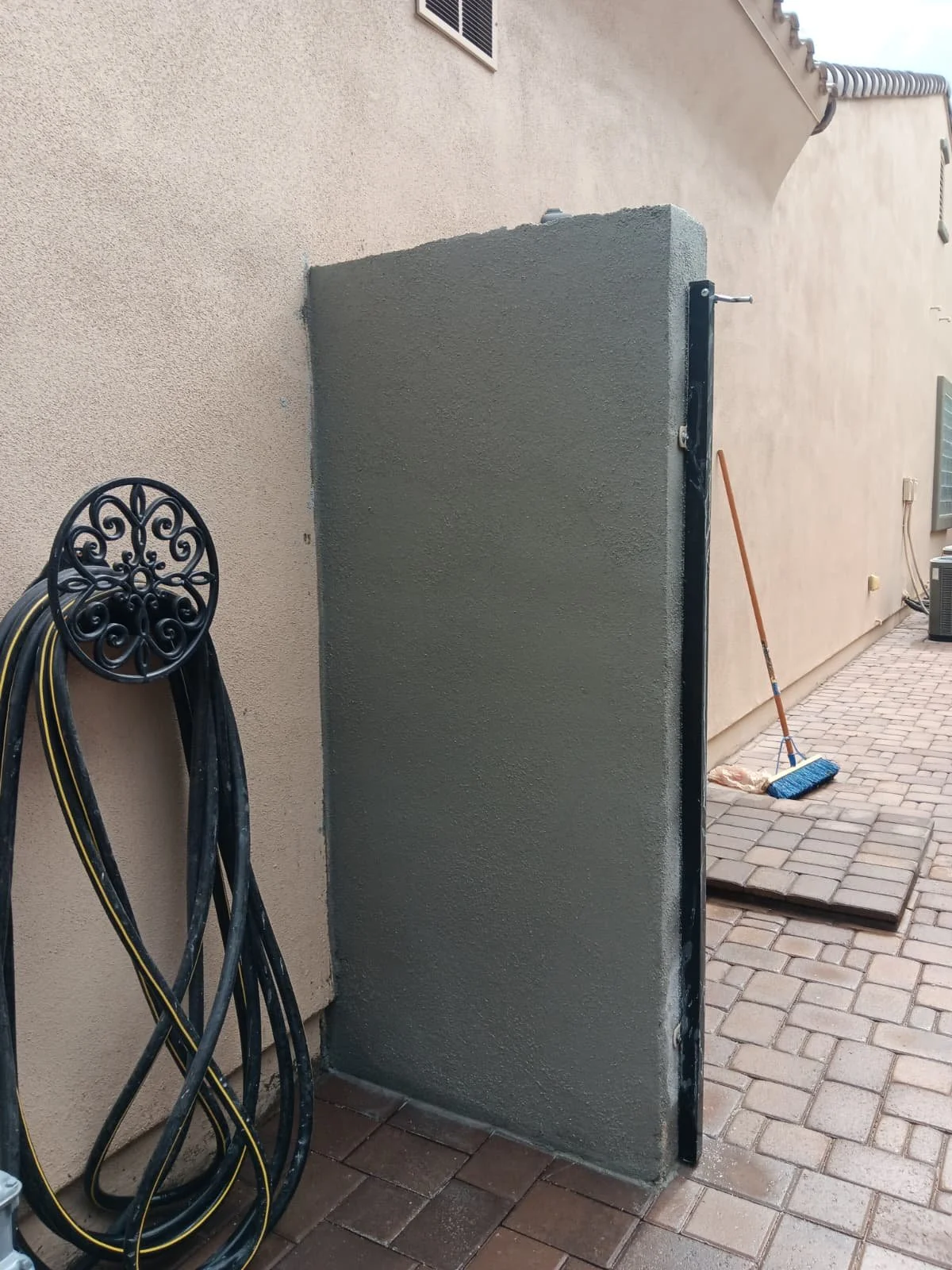 An outdoor scene showing a partially constructed or renovated wall or structure attached to a beige exterior wall of a building. There are black and yellow hoses and a decorative black metal garden ornament on the left, a broom leaning against the st