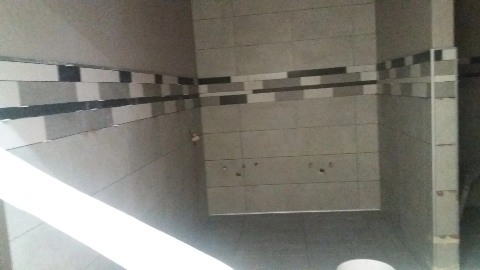 Interior of a bathroom or shower with beige and gray tiles, some of which are arranged in a pattern, and people in the reflection of a mirror.