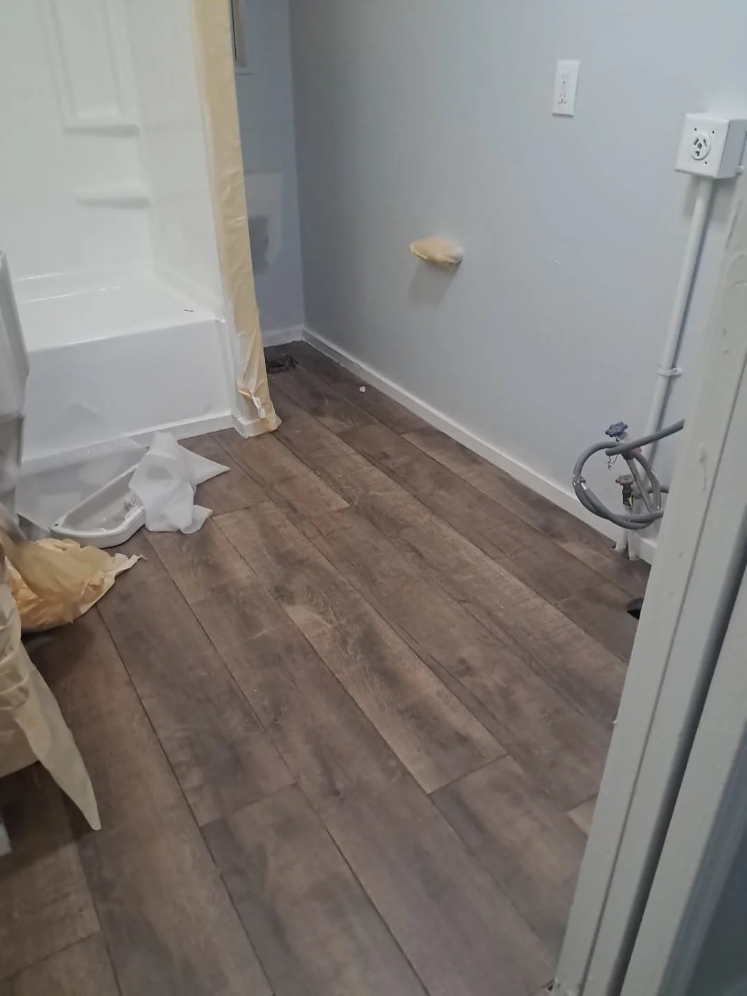 A partially renovated bathroom with a wooden floor, a shower stall to the left, and an empty wall on the right with electrical outlets and pipes. There are construction materials and supplies on the floor.