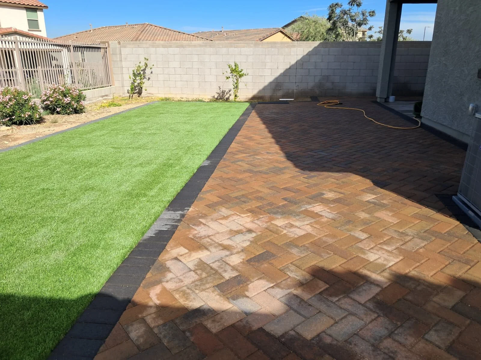 A backyard with a section of green artificial grass, a brick paved patio, a garden bed with flowering plants, and a gray concrete block fence. There is a shadow cast on part of the patio from the house roof.