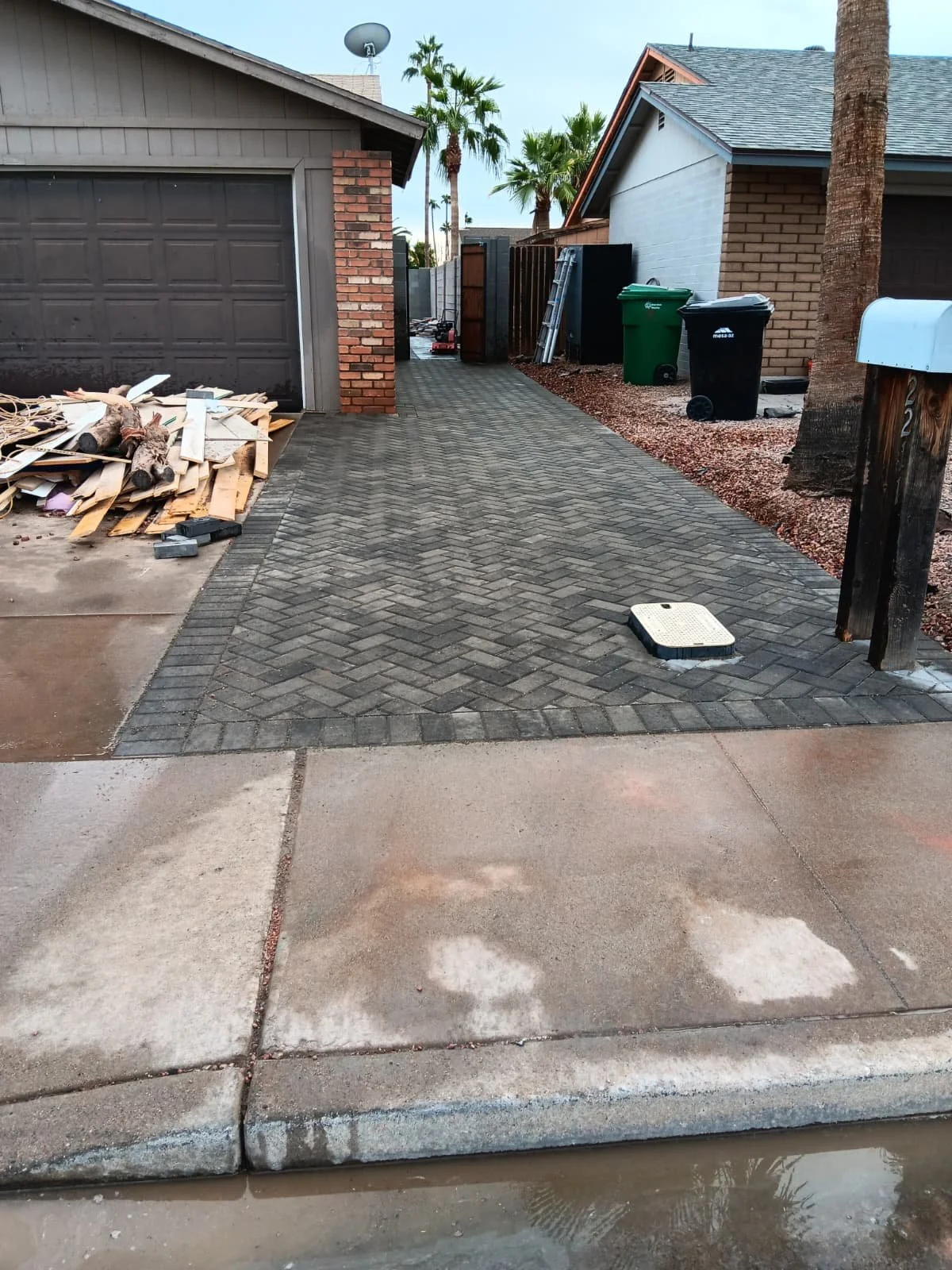 A driveway with new brick pavers leading to a garage, with a pile of discarded wood on the left, green and black trash bins, ladders, and palm trees in the background.