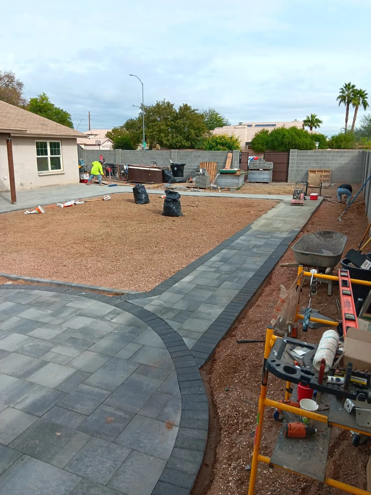 A backyard under construction with a paved pathway, a partially completed driveway, construction tools, and workers. The area is surrounded by a concrete block wall, with trees and houses visible in the background.