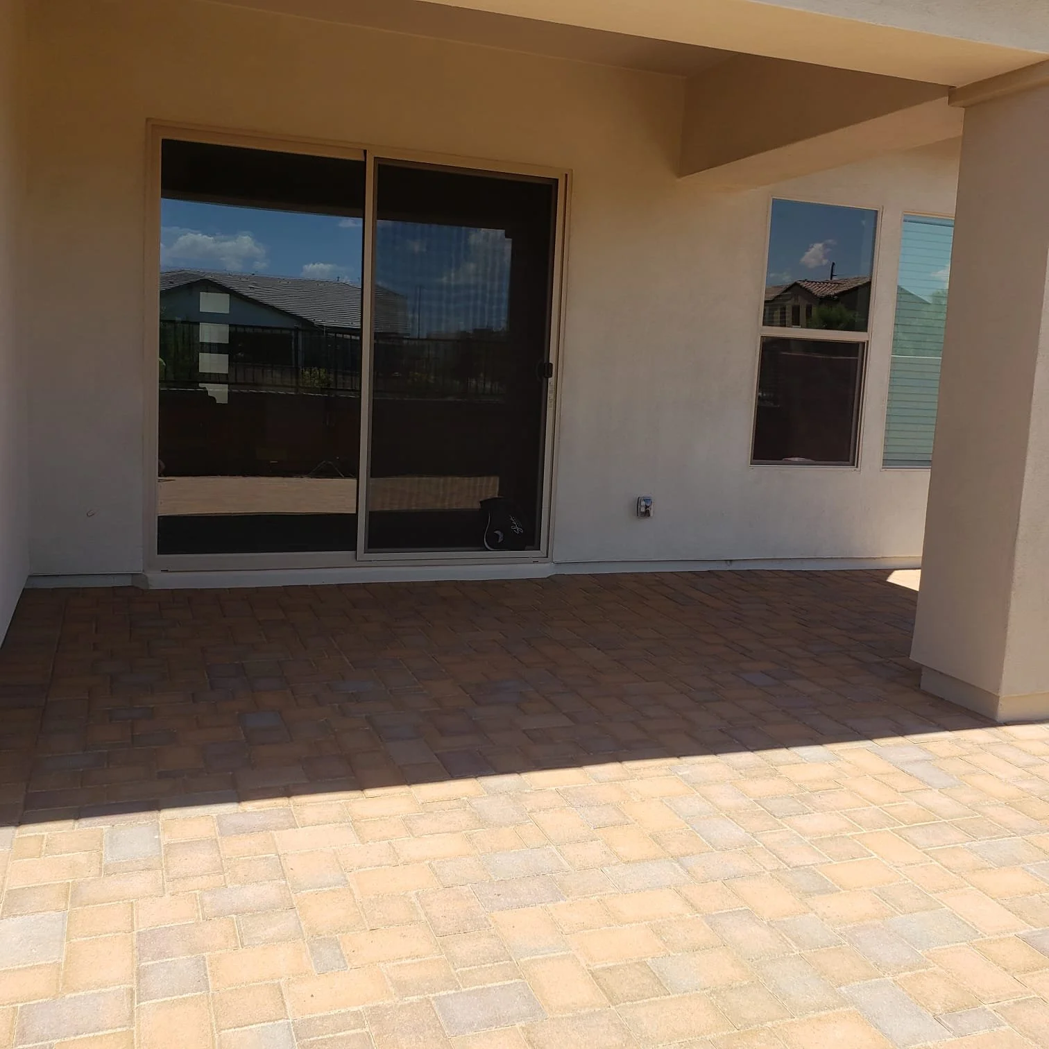 Empty covered patio with brick flooring, glass sliding door, and windows in a residential backyard.
