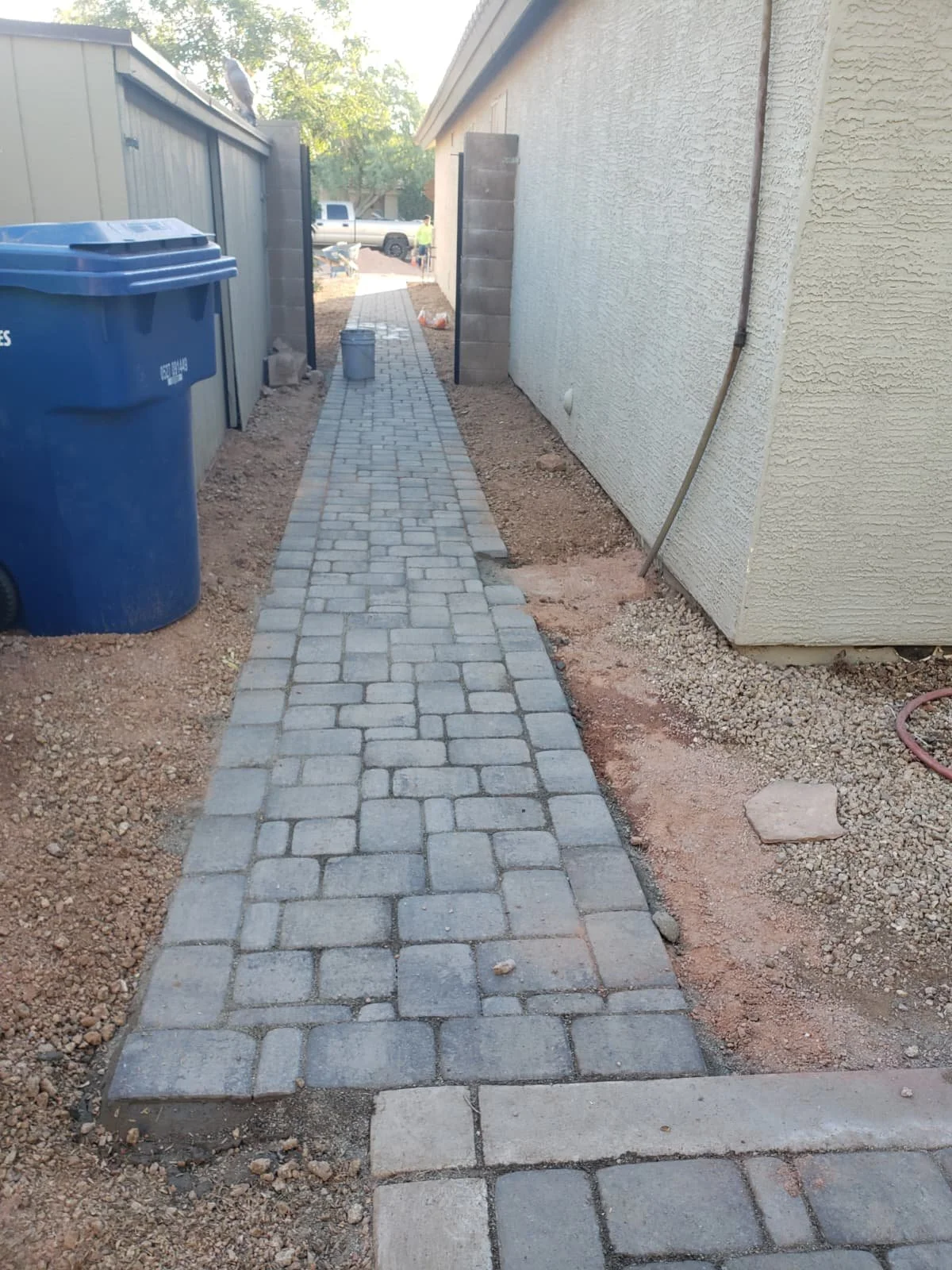 A newly paved walkway made of interlocking pavers runs between a light-colored house and a gray fence. A blue trash bin is on the left, and a bucket and construction tools are further down the walkway. There is a gate at the end of the path, leading 