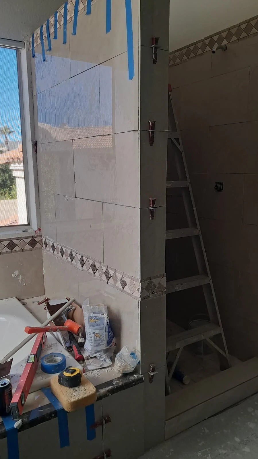 Bathroom under construction with partially tiled walls, a window, ladders, and various tools and construction supplies.