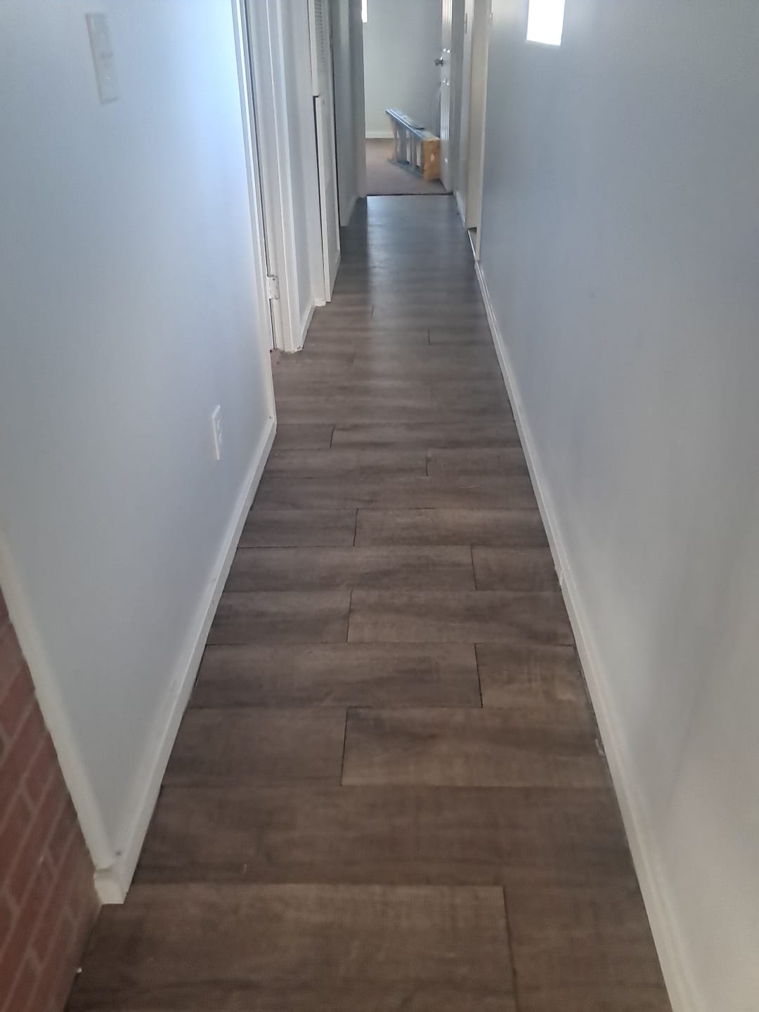 A hallway with light-colored walls, dark wood floor, and white trim, leading to a small room with a step ladder and a cardboard box visible.