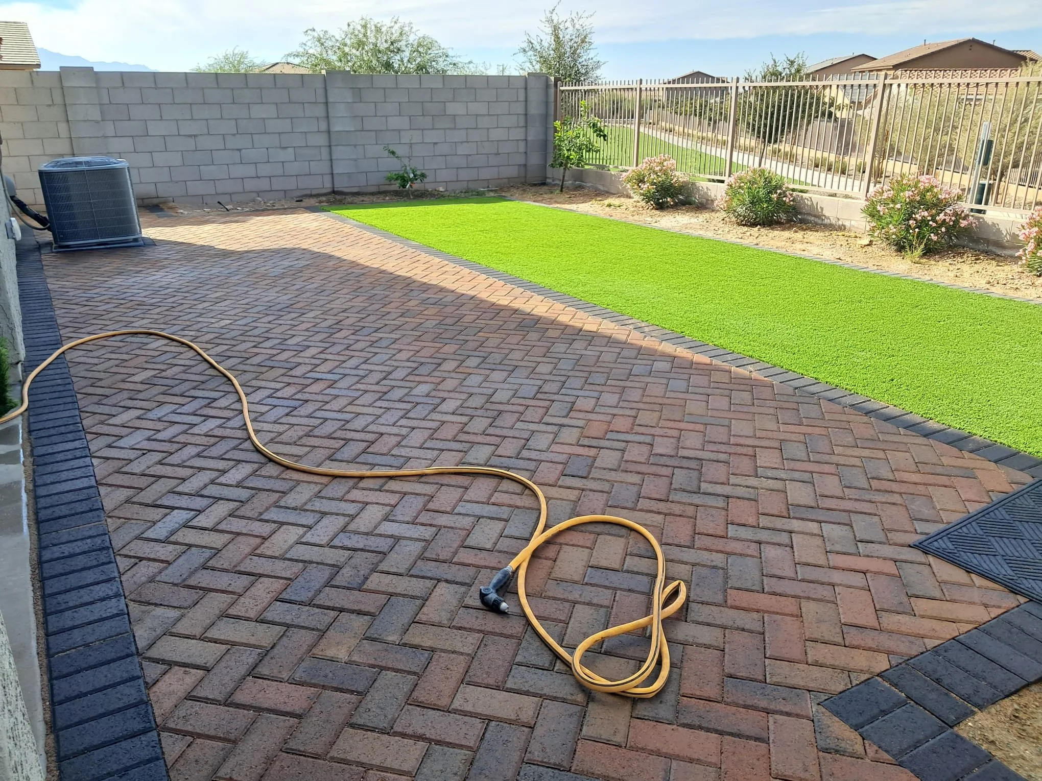 A backyard patio with a brick paver surface, a yellow garden hose resting on the ground, bordered by a concrete block wall and a metal fence, with a strip of artificial grass and flowering bushes along the fence.