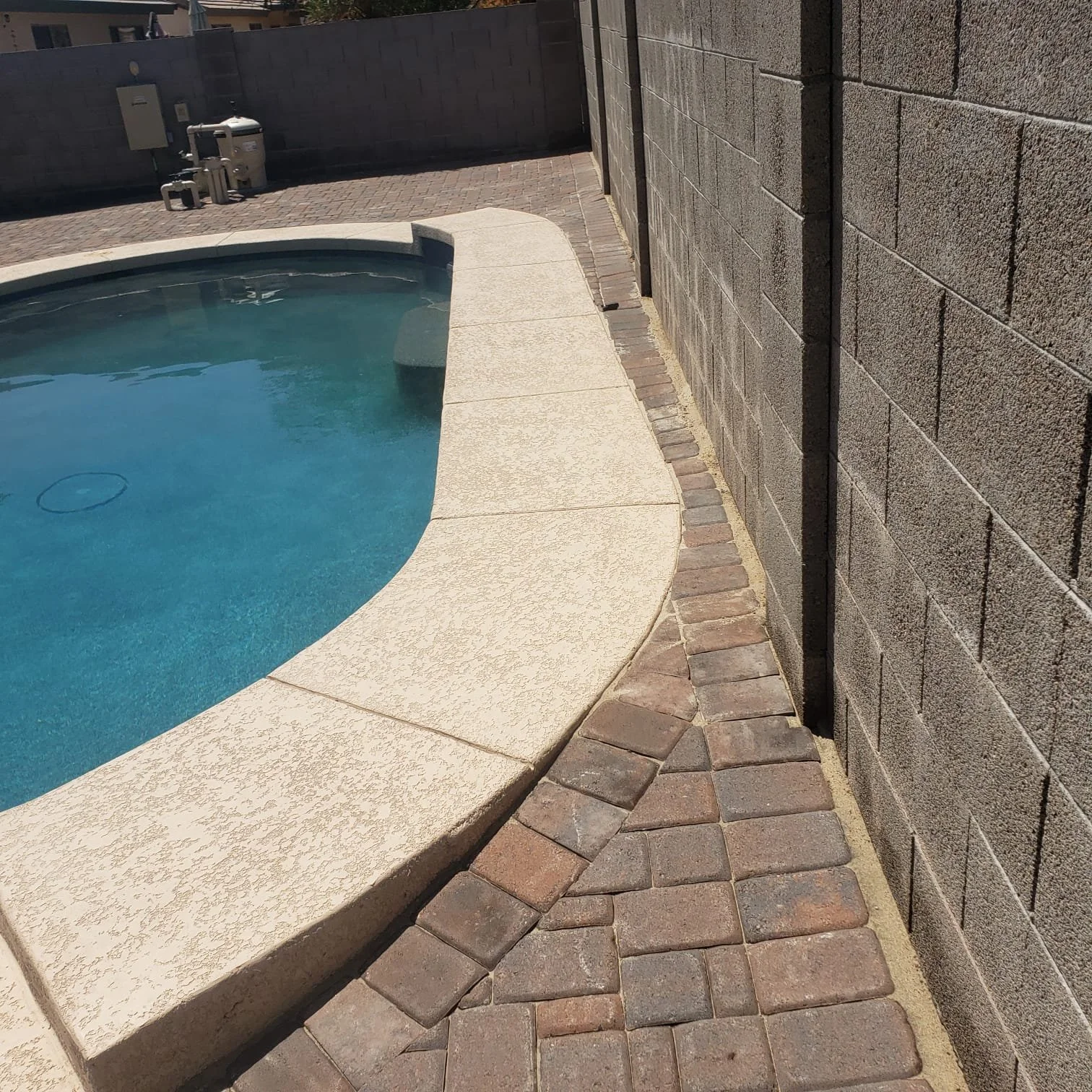 Close-up of a swimming pool edge with beige textured concrete and brick paving, next to a concrete block wall under bright sunlight.