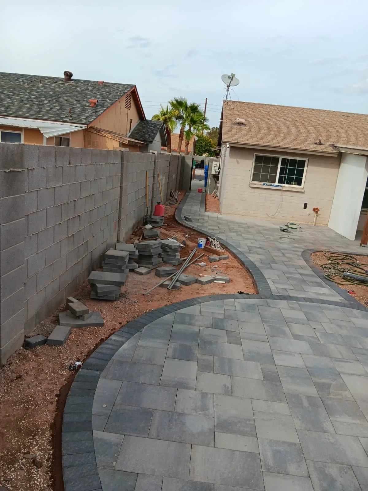 A backyard under construction with a new paved patio, a curved pathway, some construction materials, and tools. A wall with trees behind it is on the left, and there are houses with satellite dishes and palm trees in the background.