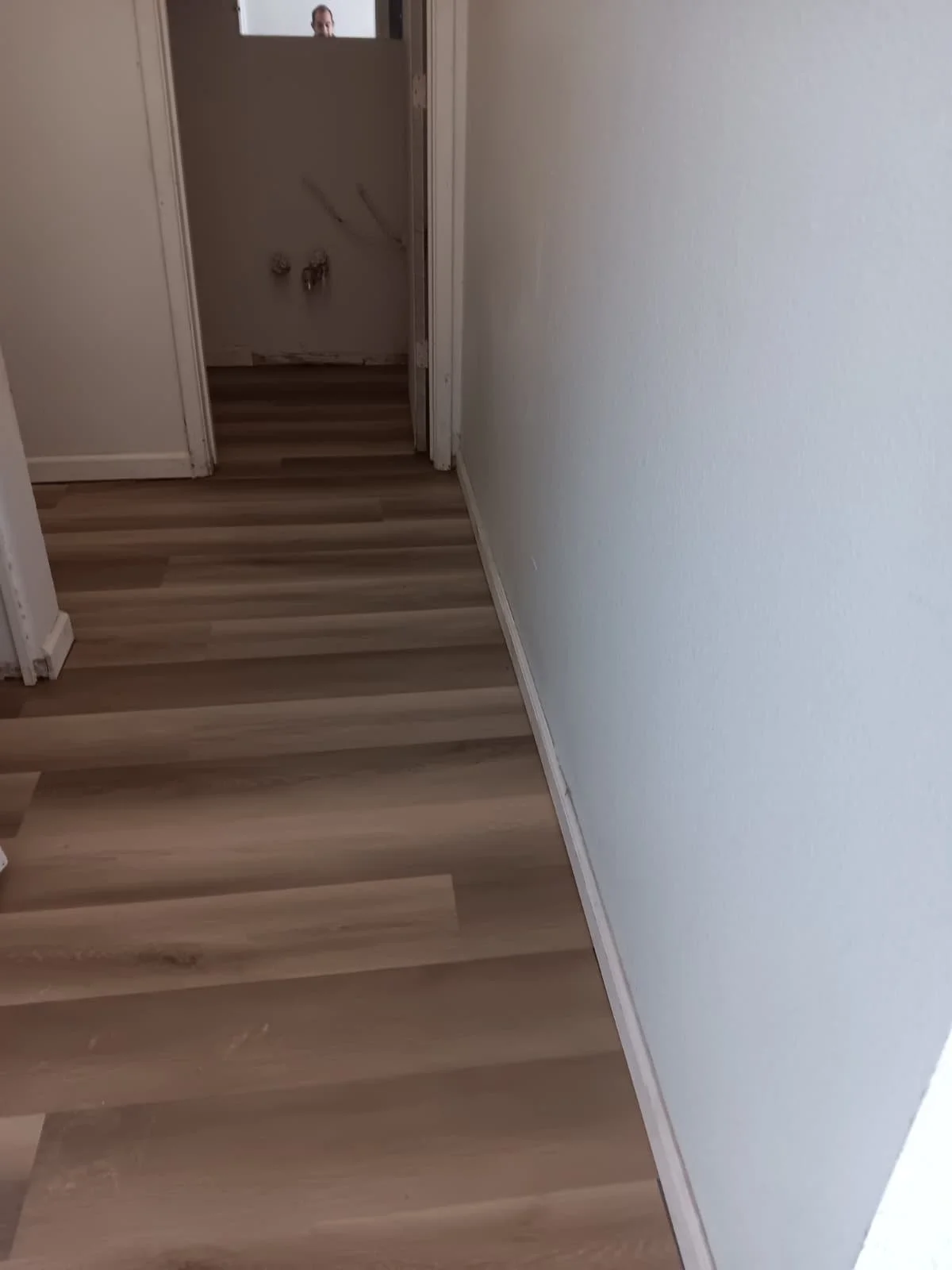 Empty hallway with wood-patterned flooring and plain white walls, leading to a room under renovation with exposed pipes and drywall.