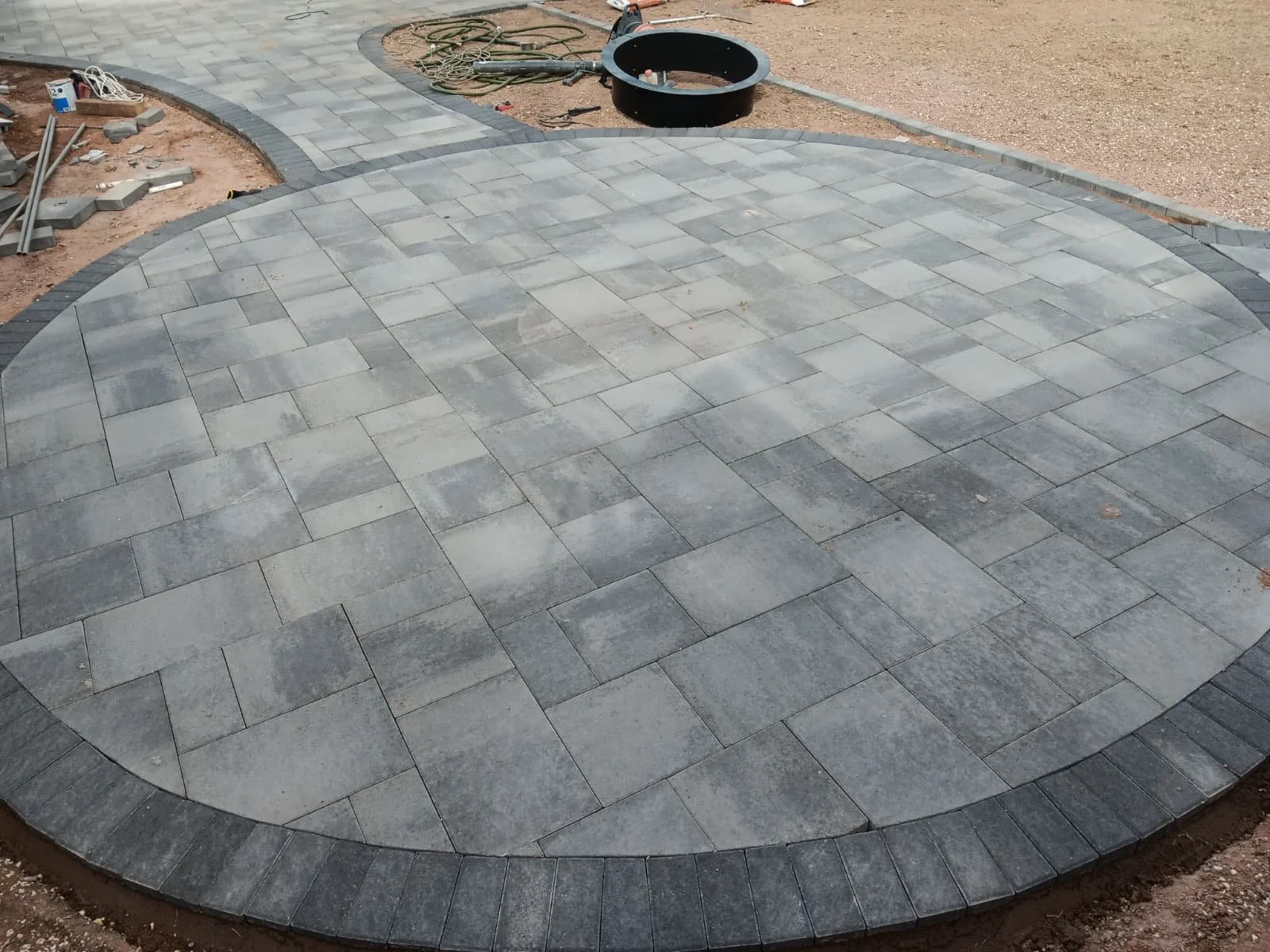 Gray stone pavers arranged in a curved pattern, forming a small patio or walkway under construction, with construction tools and materials scattered nearby.