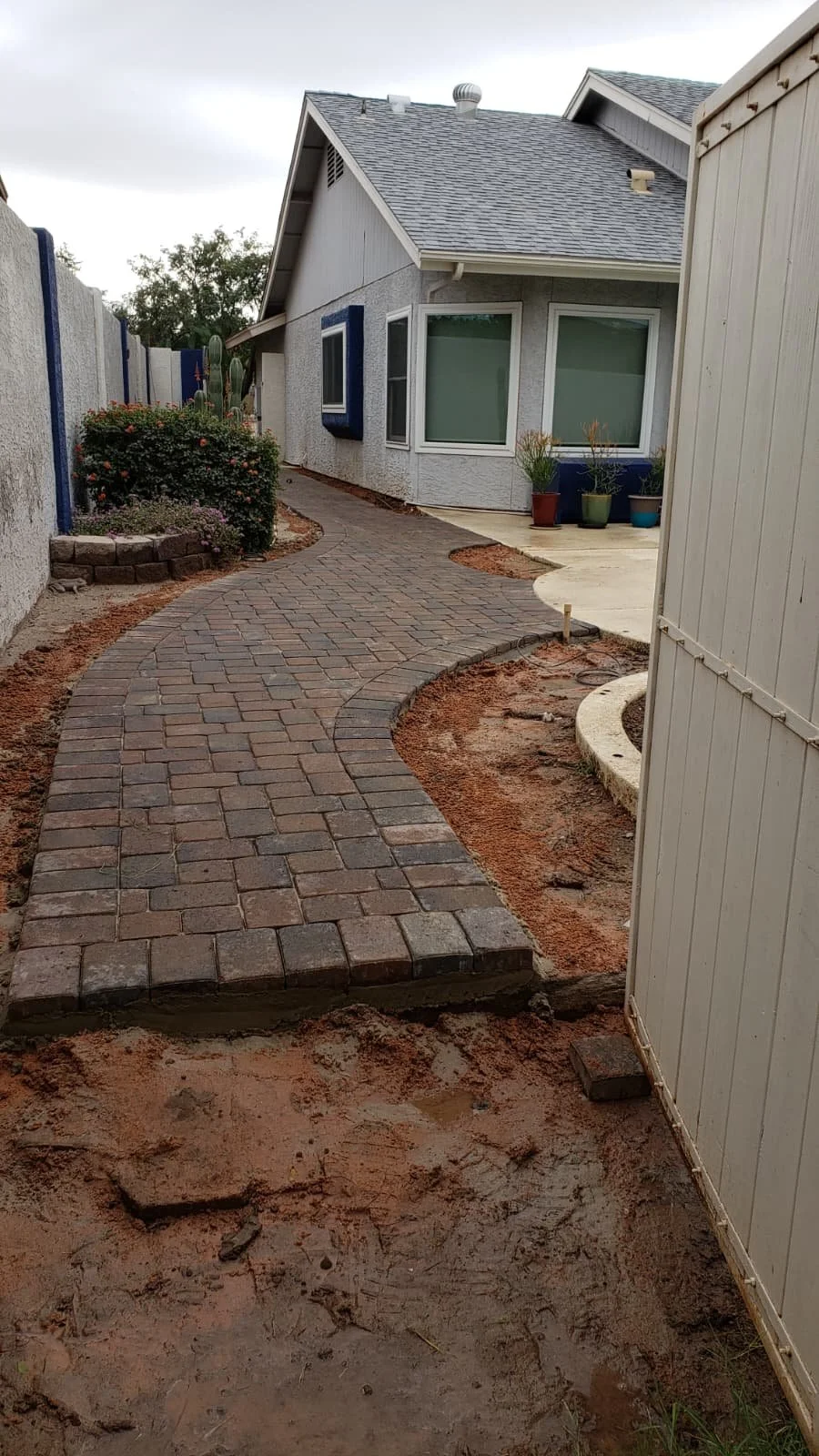 Curved brick pathway leading to a house with gray walls and large windows, with potted plants next to the house and a flower bed with bushes and cactus on the left.