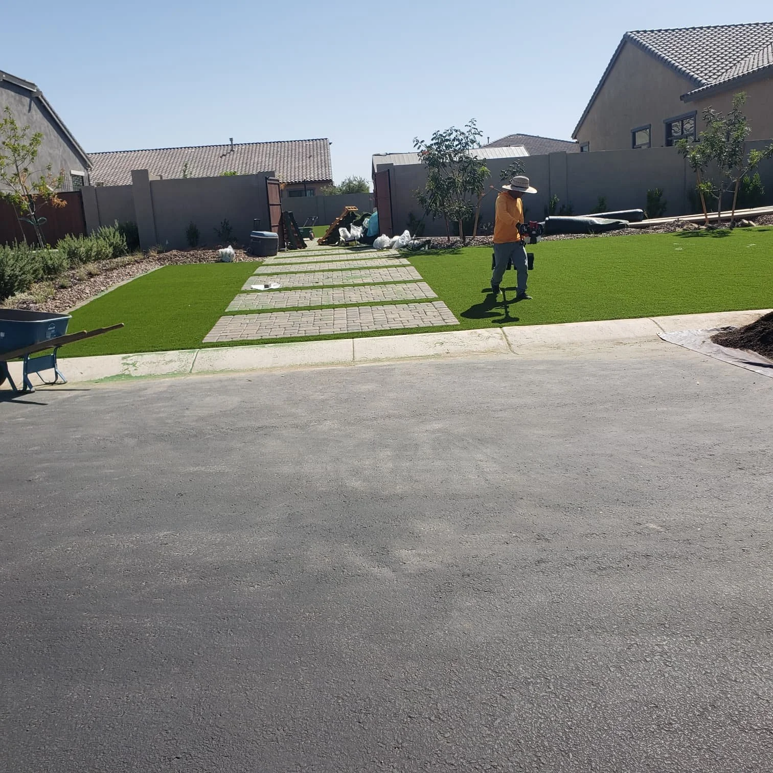 A person working on installing or maintaining a lawn in a residential backyard, with a paved pathway, trees, and houses in the background.