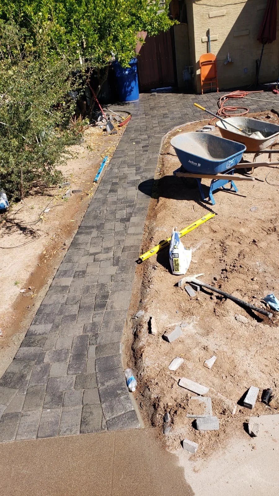 A newly paved walkway under construction in a backyard, with wheelbarrows, tools, and bricks scattered around.