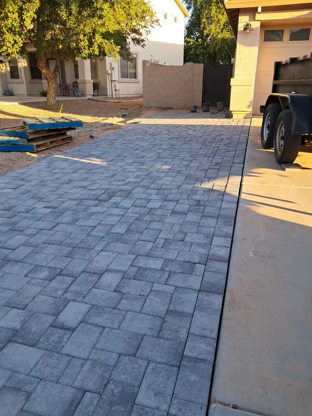 Newly paved stone driveway with some construction materials and truck parked beside it, in a backyard with trees, a small brick wall, and neighboring houses in the background.