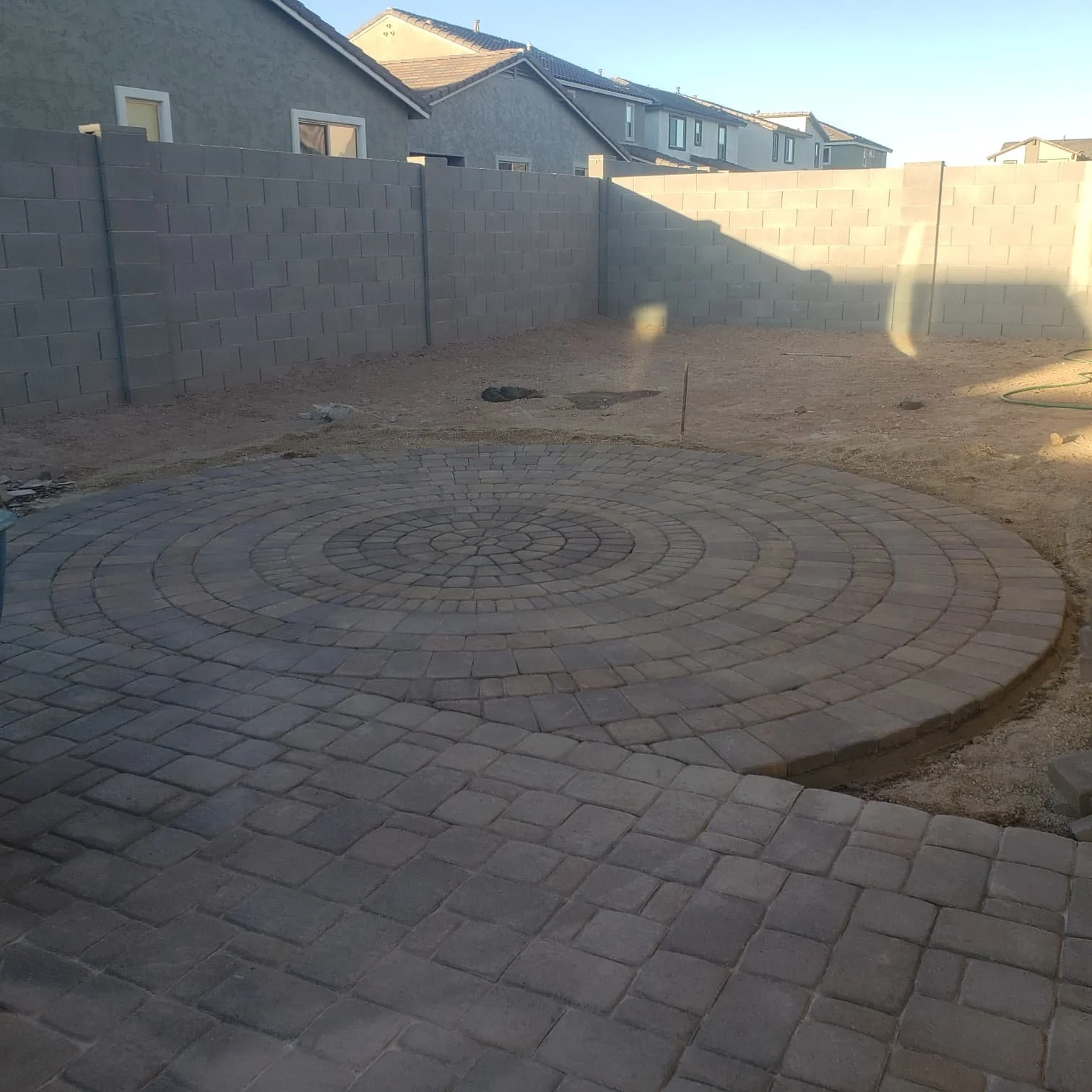 A backyard with a circular brick patio under construction, surrounded by a dirt area with a gray concrete block wall, neighboring houses, and a clear sky in the background.