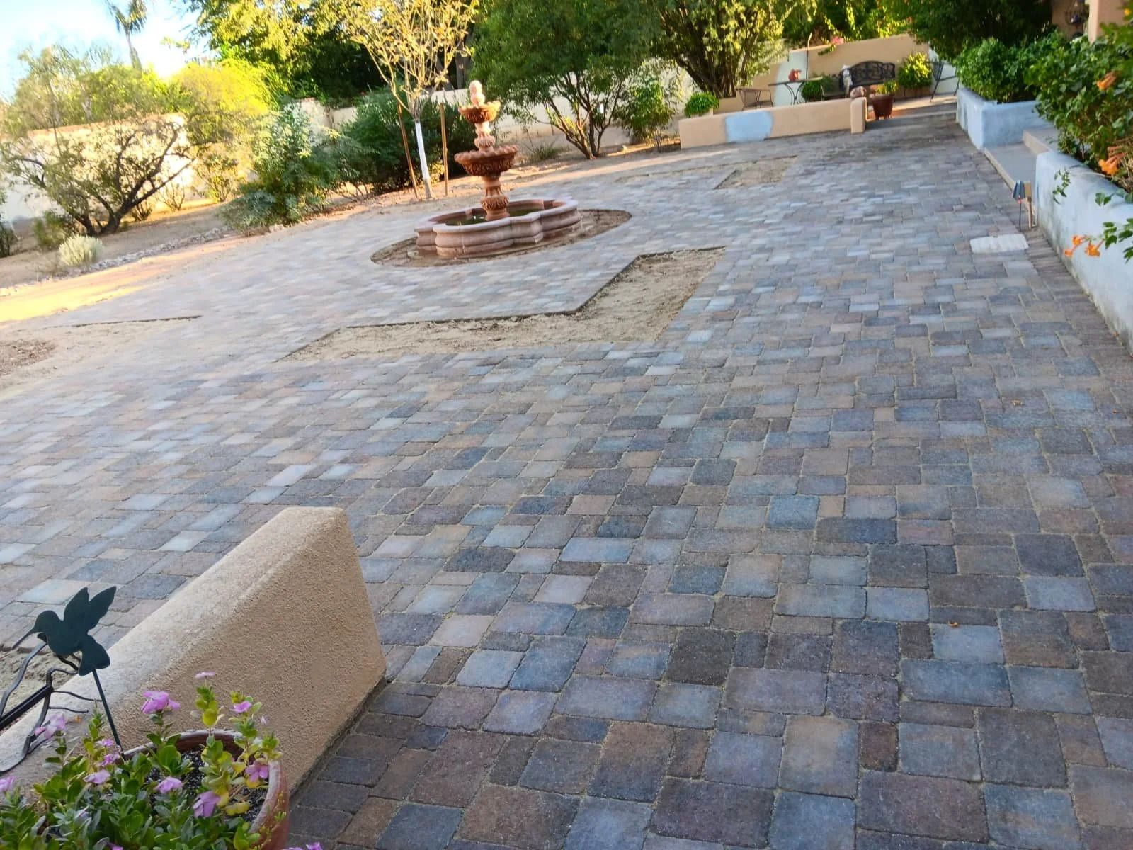 A paved outdoor courtyard with a fountain, benches, trees, and potted plants.