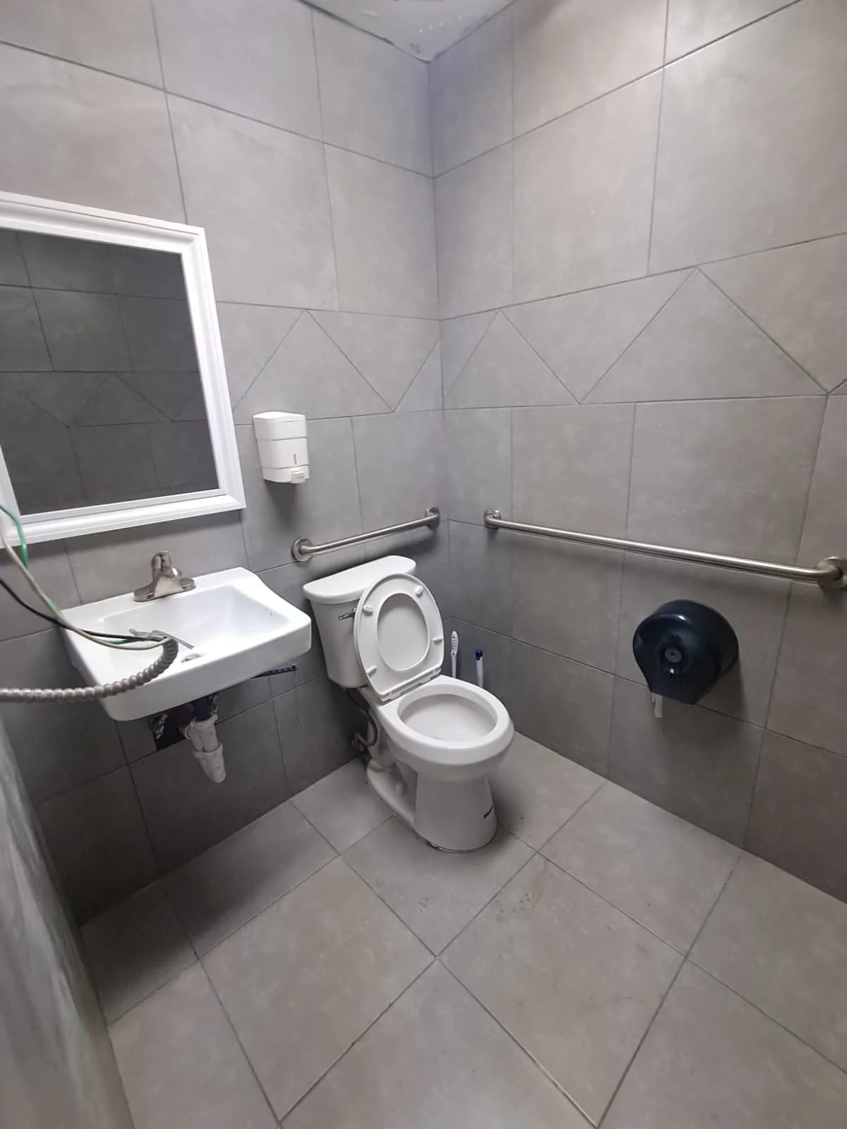 A small, gray-tiled accessible restroom with a toilet, a white wall-mounted sink, a mirror, a soap dispenser, and grab bars on the walls.