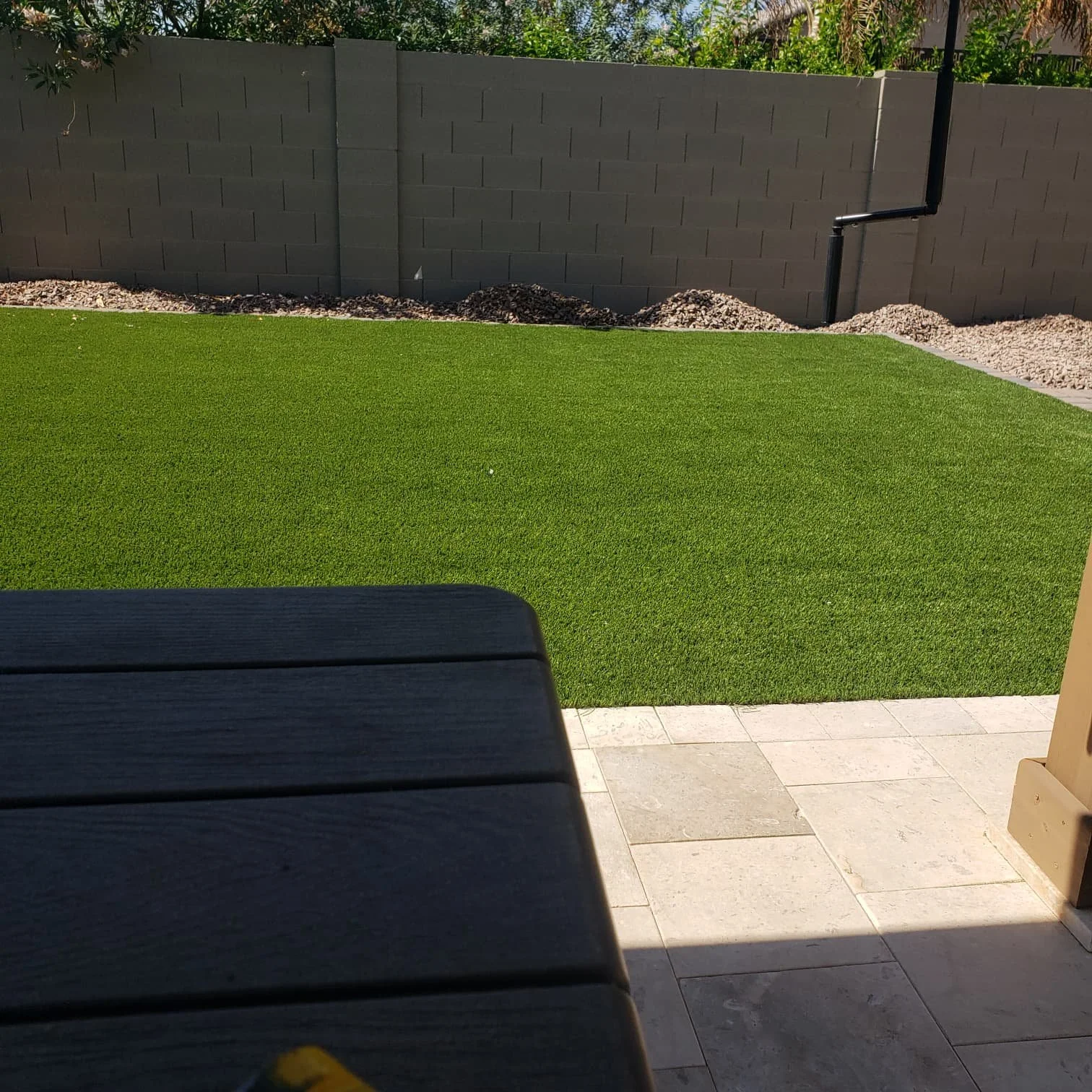 View of a backyard with a green artificial turf lawn, a concrete stone patio, and a gray cinder block wall with a black drain pipe running down it.