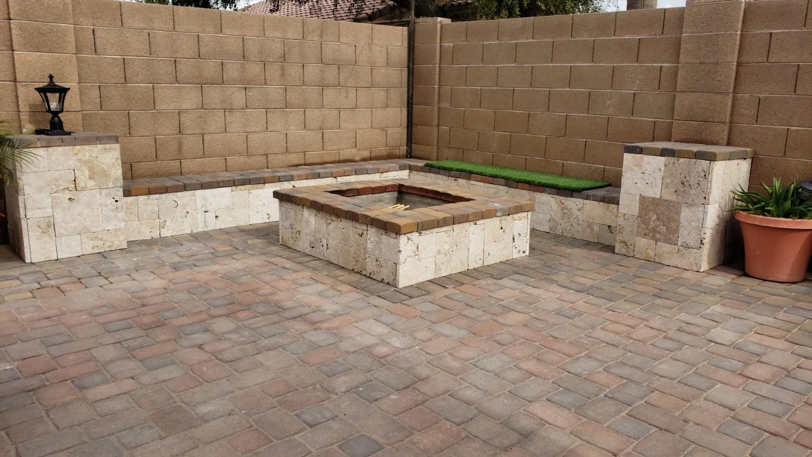 An outdoor patio with brick pavers, built-in seating, a fire pit in the center, and decorative planter with plant on the right, surrounded by a brick wall.