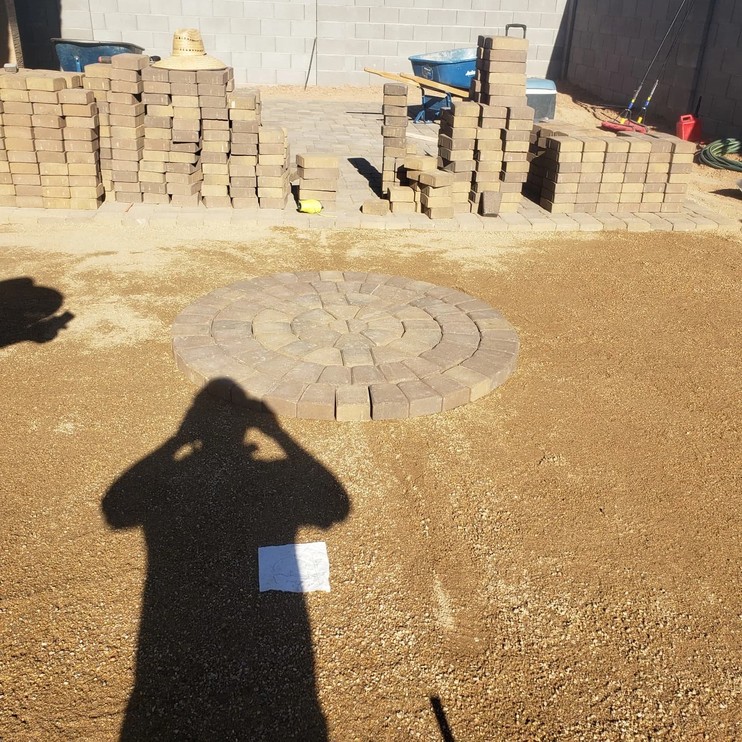 This image shows a backyard with a newly built circular brick patio at the center, surrounded by dirt. In the background, there are stacks of bricks, tools, and construction supplies, indicating ongoing landscaping or patio work.