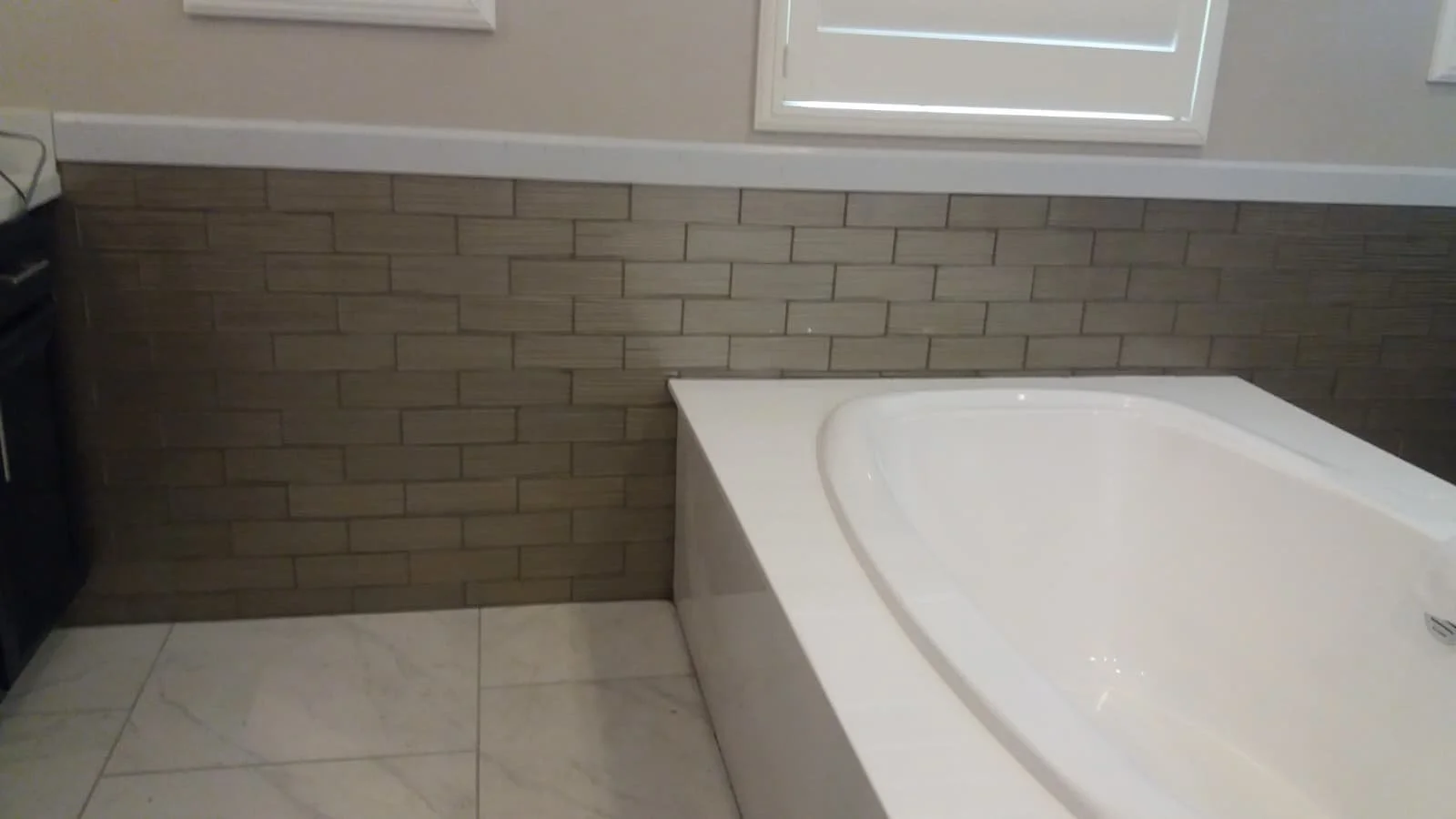 Bathroom with a large white bathtub, brown brick-patterned wall, beige floor tiles, and a window above the bathtub.