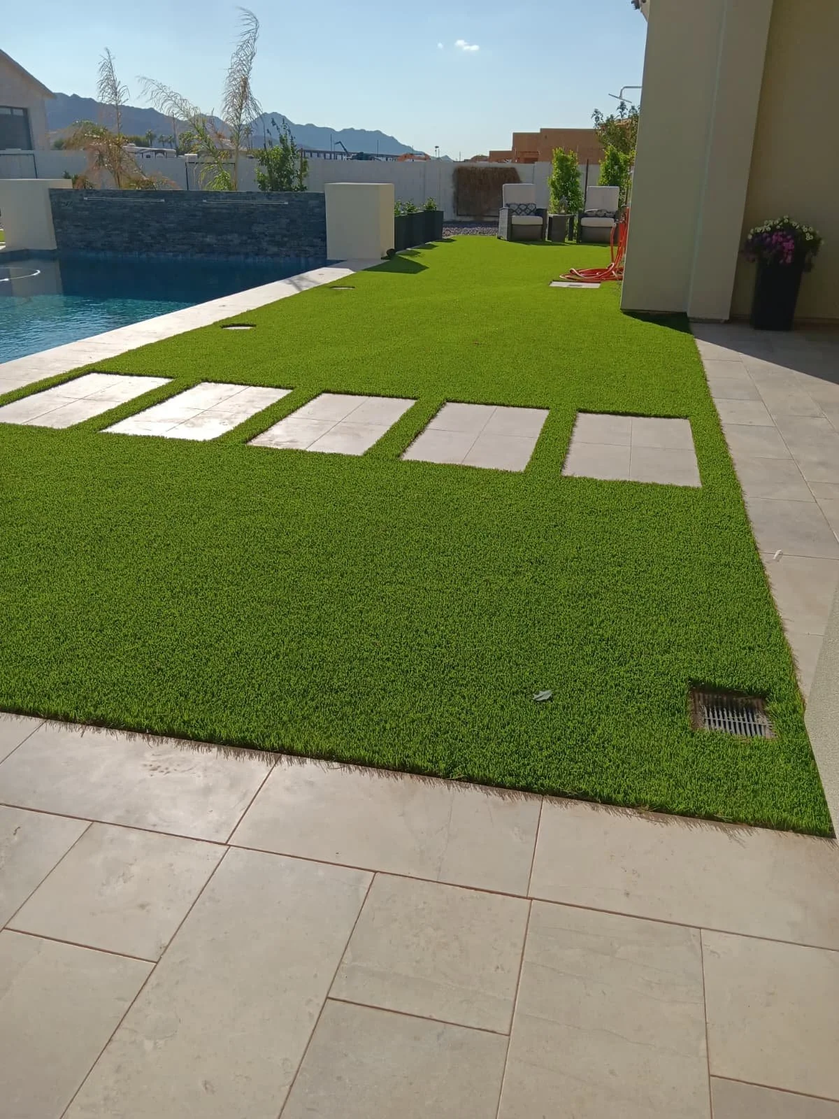 Backyard with green artificial turf, partially bordered by beige tiles, and some decorative square paving stones. There is a swimming pool on the left, a few patio chairs, and mountain scenery in the background.