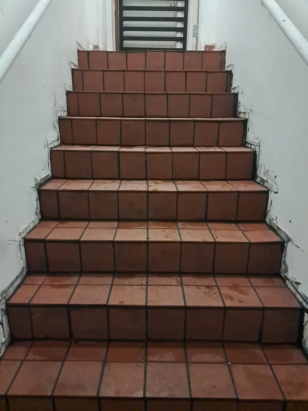 Indoor tiled staircase under construction with incomplete white wall paint and visible wall damage on sides.