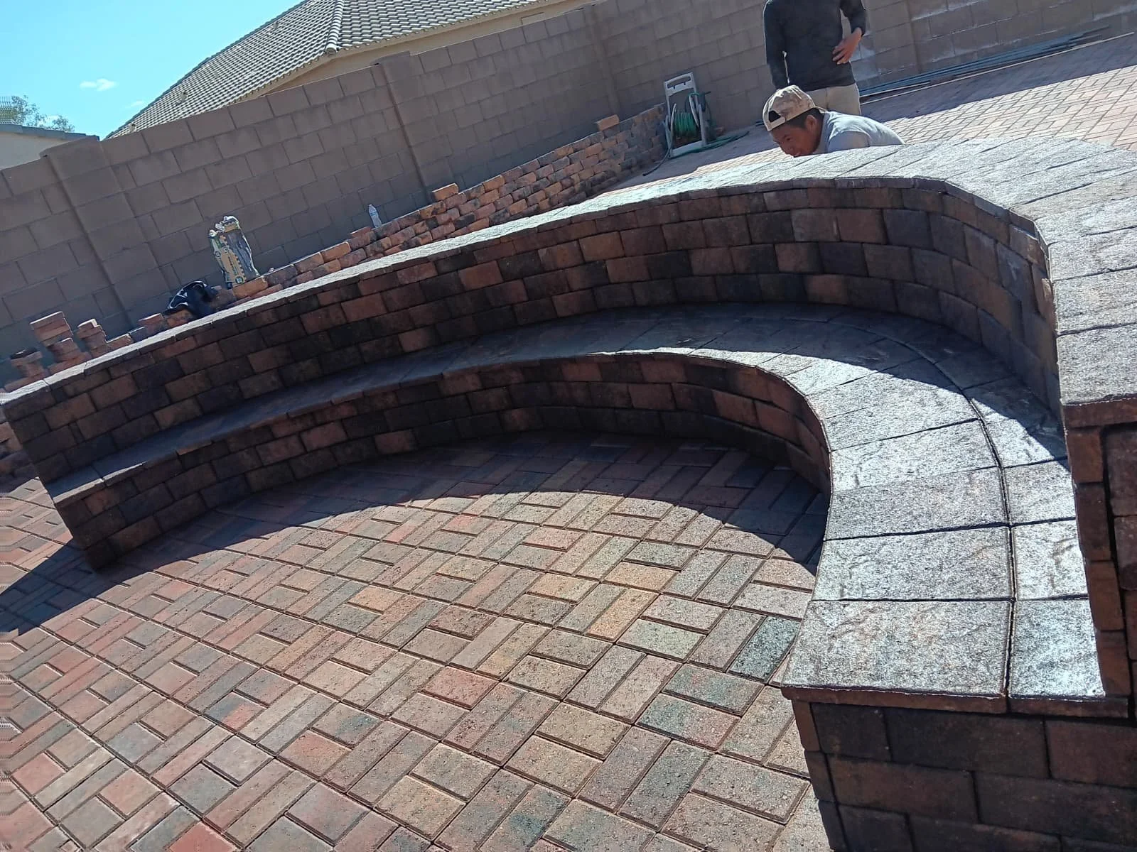 Person laying on a brick curved bench in a backyard with another person standing nearby, building a brick wall, and outdoor tools visible in the background.
