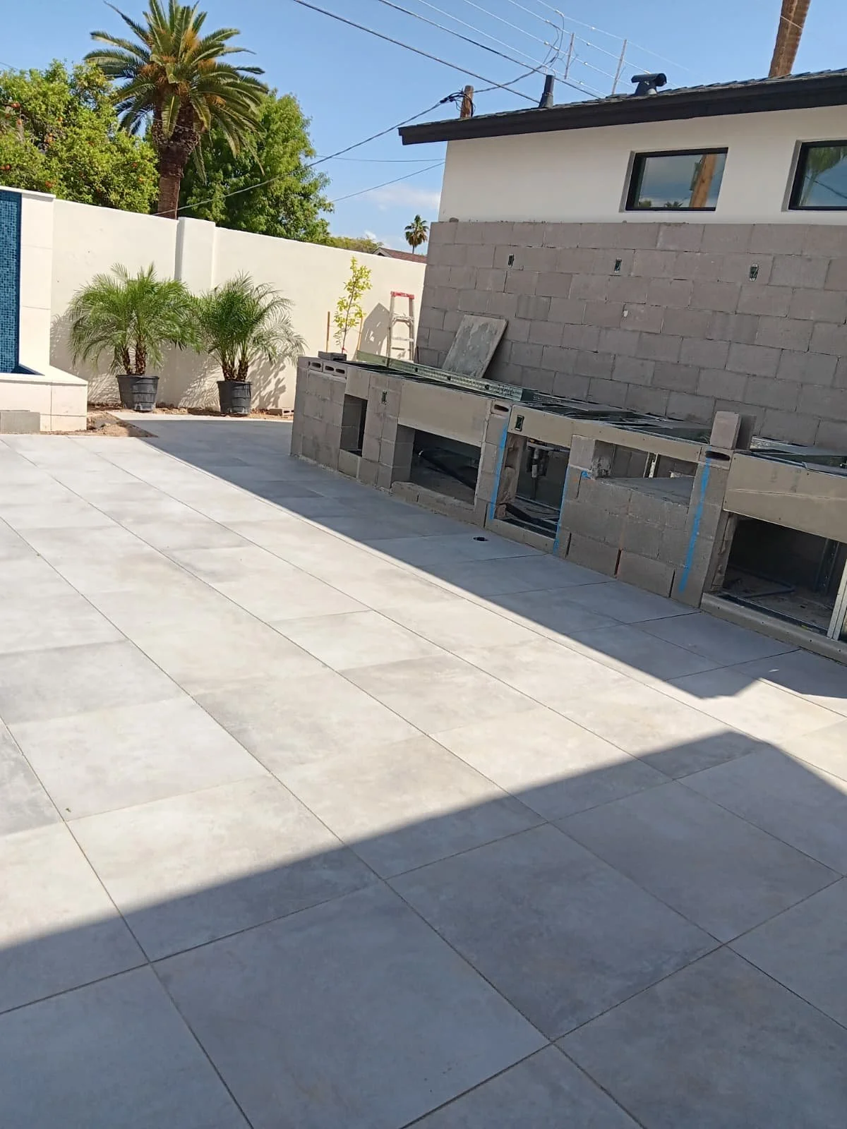 A backyard patio with tiled flooring, potted palm plants along the white wall to the left, and an unfinished outdoor kitchen with concrete block counter and built-in fixtures under construction. There is a ladder and construction materials nearby, an