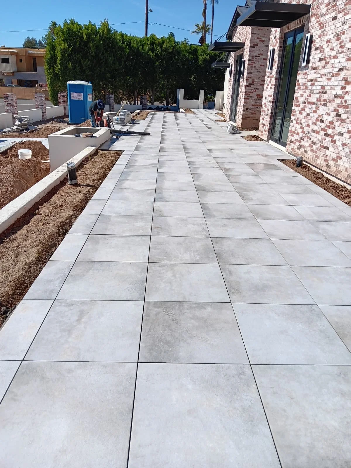 Construction workers are installing large, light-colored tiles on a patio outside a brick building. The area is still under construction, with exposed soil and construction materials visible. A blue portable toilet and some landscaping elements are p