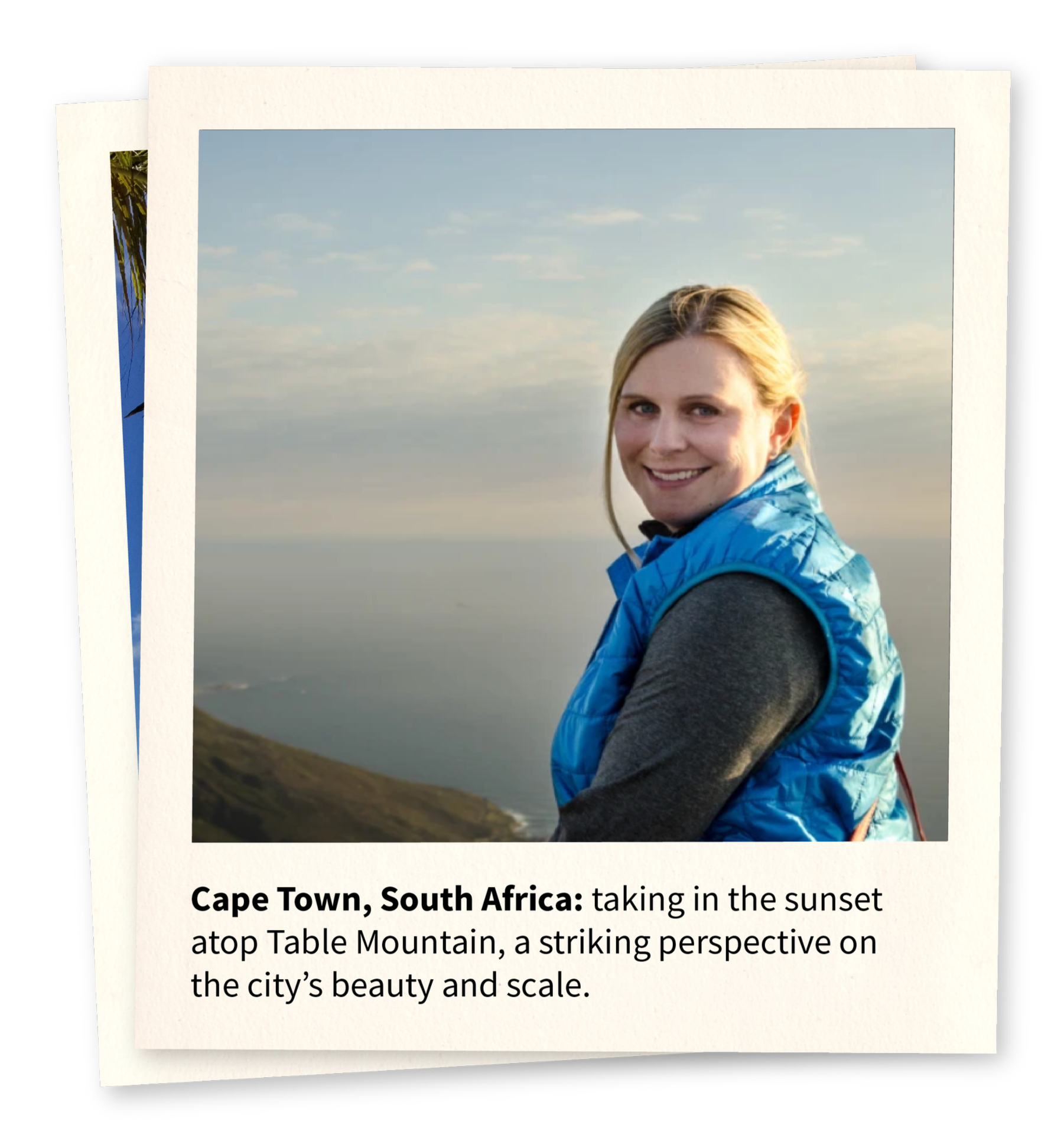 A woman smiling outdoors with a view of the ocean and a cloudy sky in Cape Town, South Africa, wearing a blue vest over a gray long sleeve shirt.