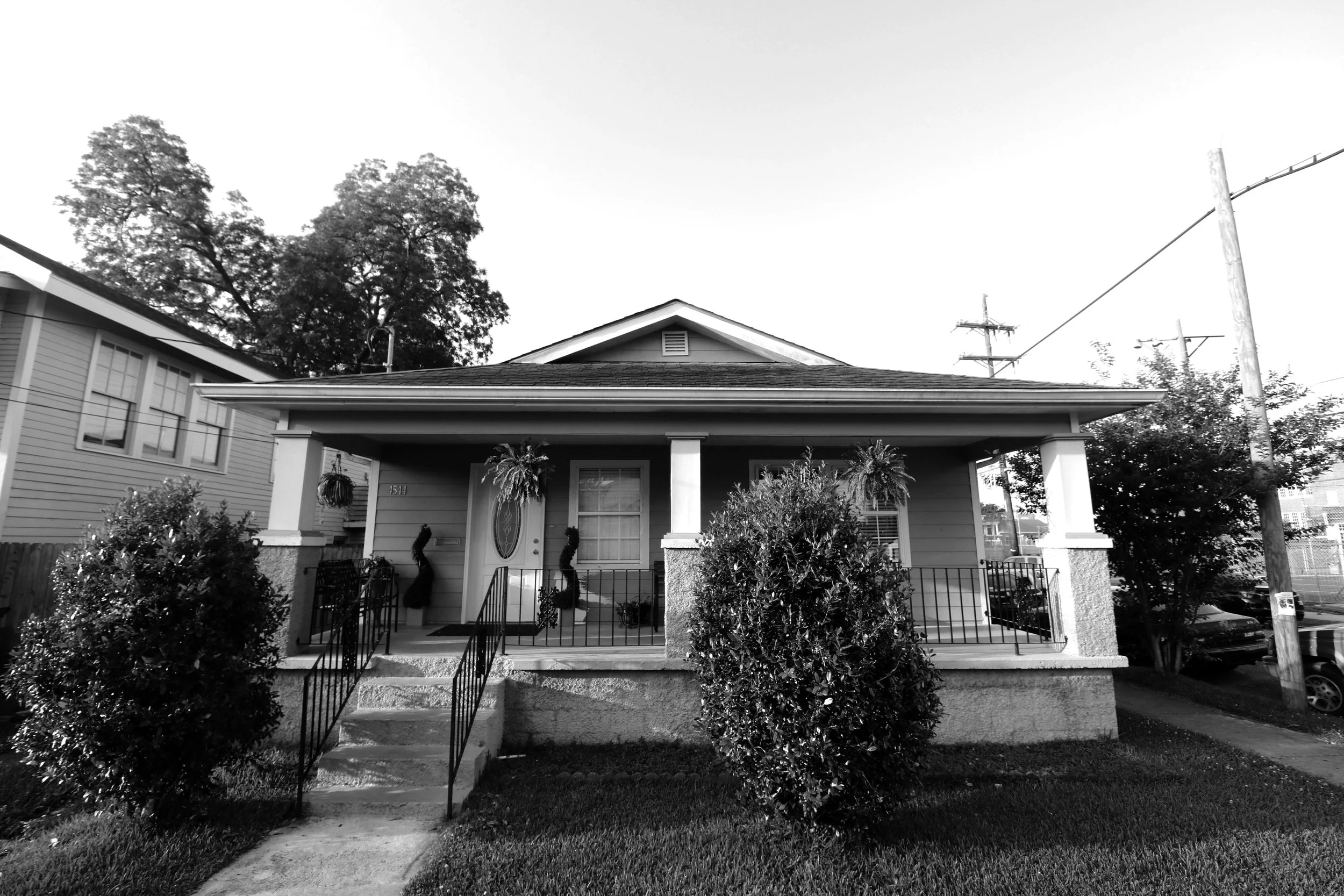 Black and white image of a single-story house with a front porch, steps leading up to it, and two large bushes in front, with hanging plants and decorative wreaths near the front door.