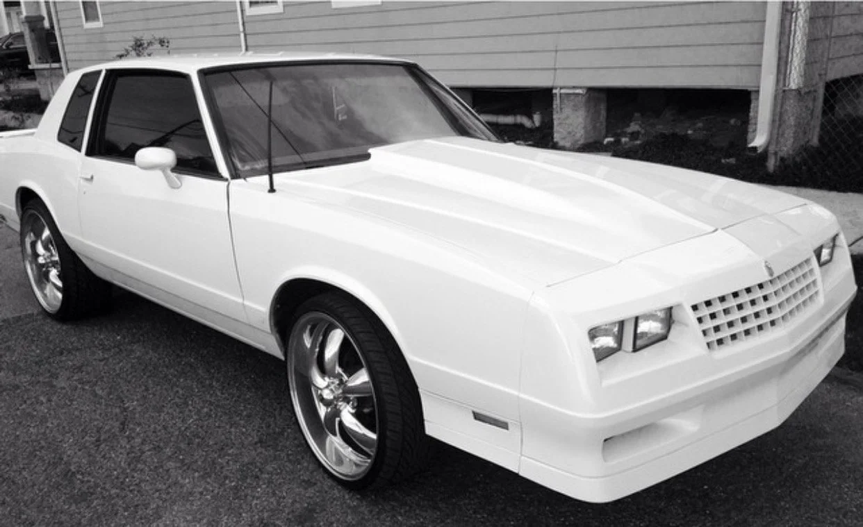 A white vintage sports car parked on the street in front of a house with siding, featuring alloy wheels and tinted windows.