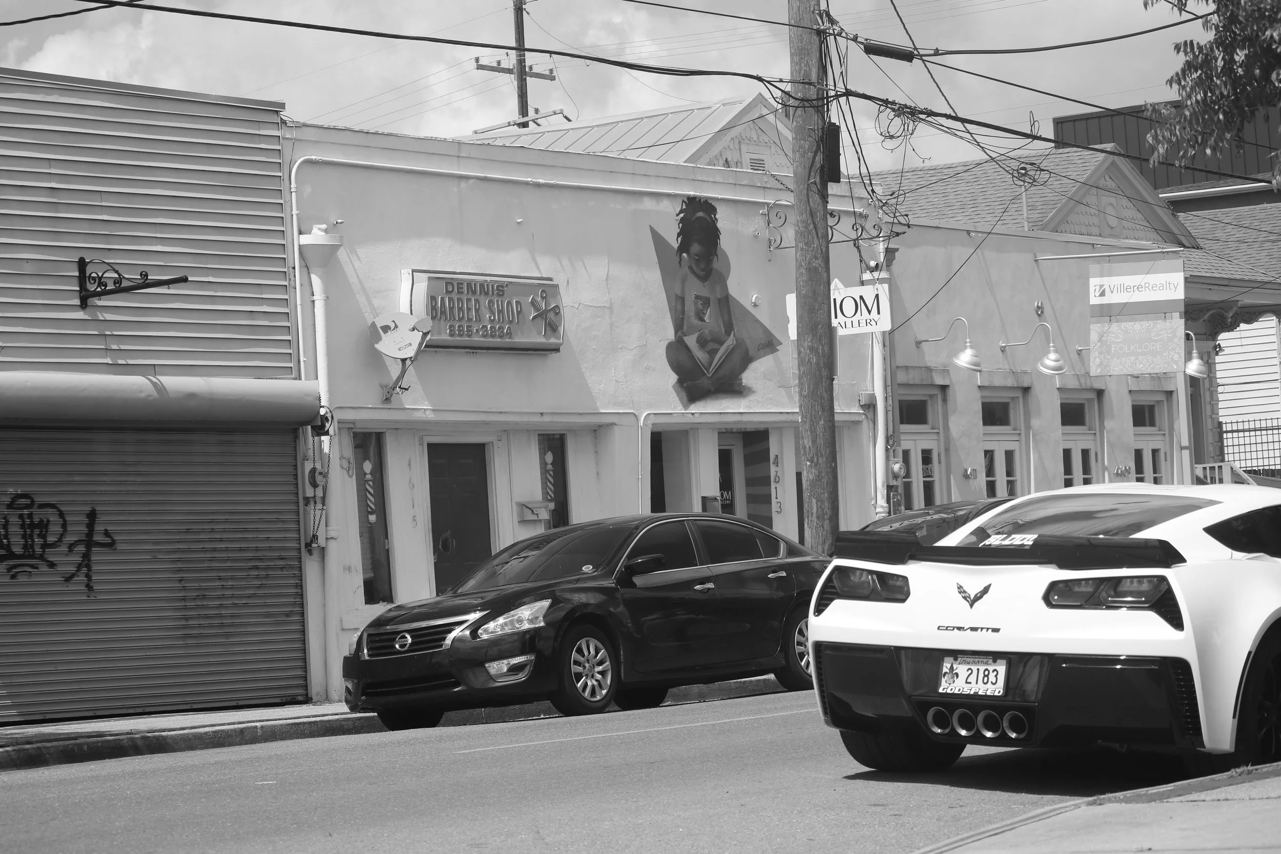 Street scene with parked cars and storefronts, including a barber shop, an art gallery, and a realty sign.