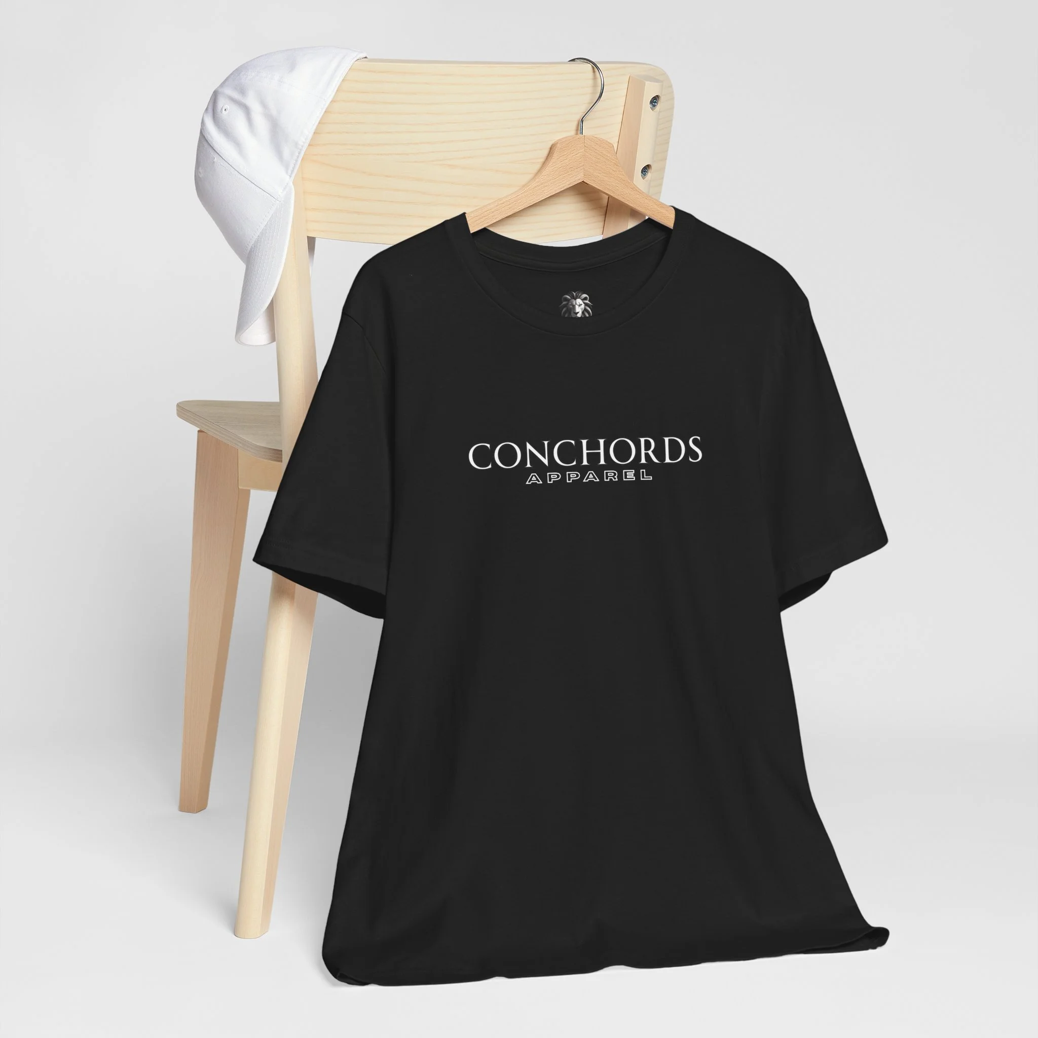 Black t-shirt hanging on wooden hanger with 'CONCHORDS APPAREL' logo, white cap on wooden chair, white background.