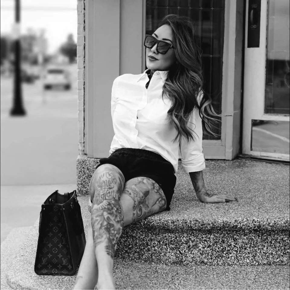 A woman with long hair, wearing sunglasses, a white shirt, and shorts, sitting on a sidewalk ledge outside a building with a bag beside her.